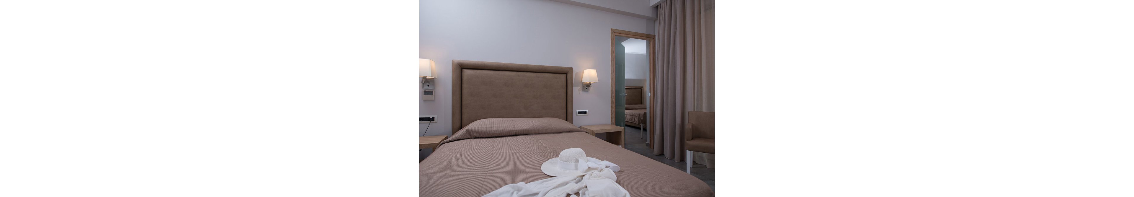 A contemporary hotel room features a neatly made bed with taupe bedding and a padded headboard. A white sunhat and light clothing are placed on the bed, adding a casual touch. The room is softly lit by wall-mounted lamps, and a mirror reflects a similar setup in an adjacent space. The palette is neutral, with beige and brown tones creating a calm, inviting atmosphere.