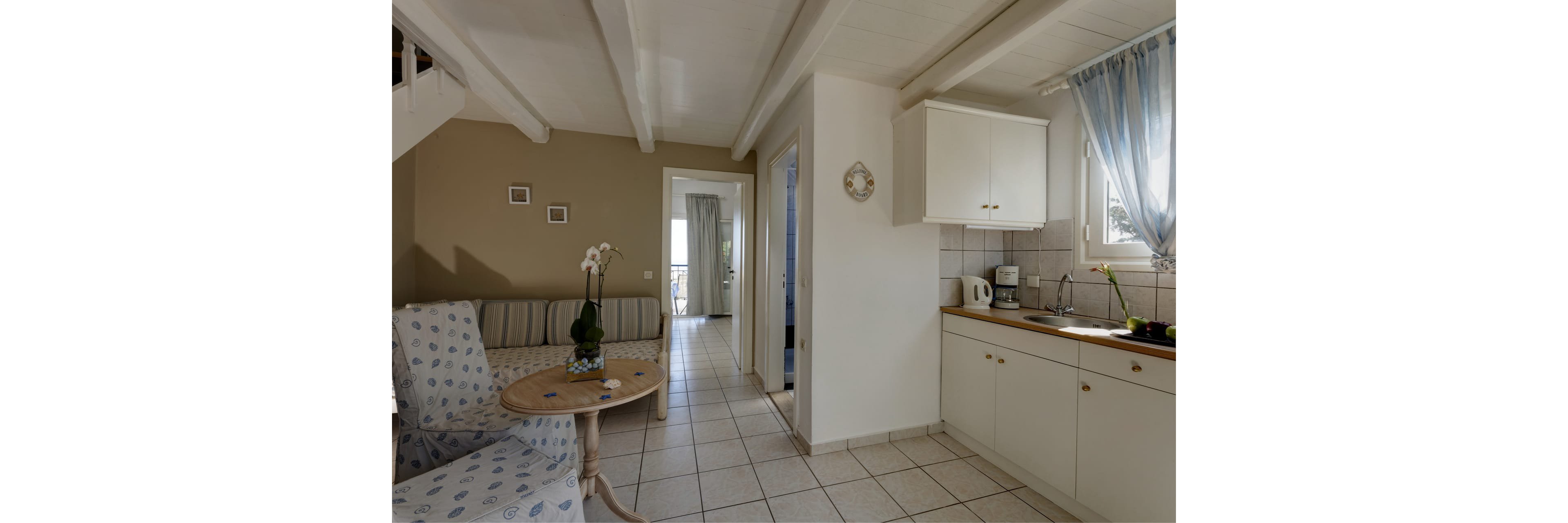 Bright, compact kitchen opens into a small living area with a patterned armchair and striped sofa. White cabinets, tiled backsplash, and countertop appliances create a simple, functional cooking space. A round wooden table with a potted orchid adds a decorative touch near the seating. Light curtains and neutral walls give the room an airy, relaxed atmosphere.