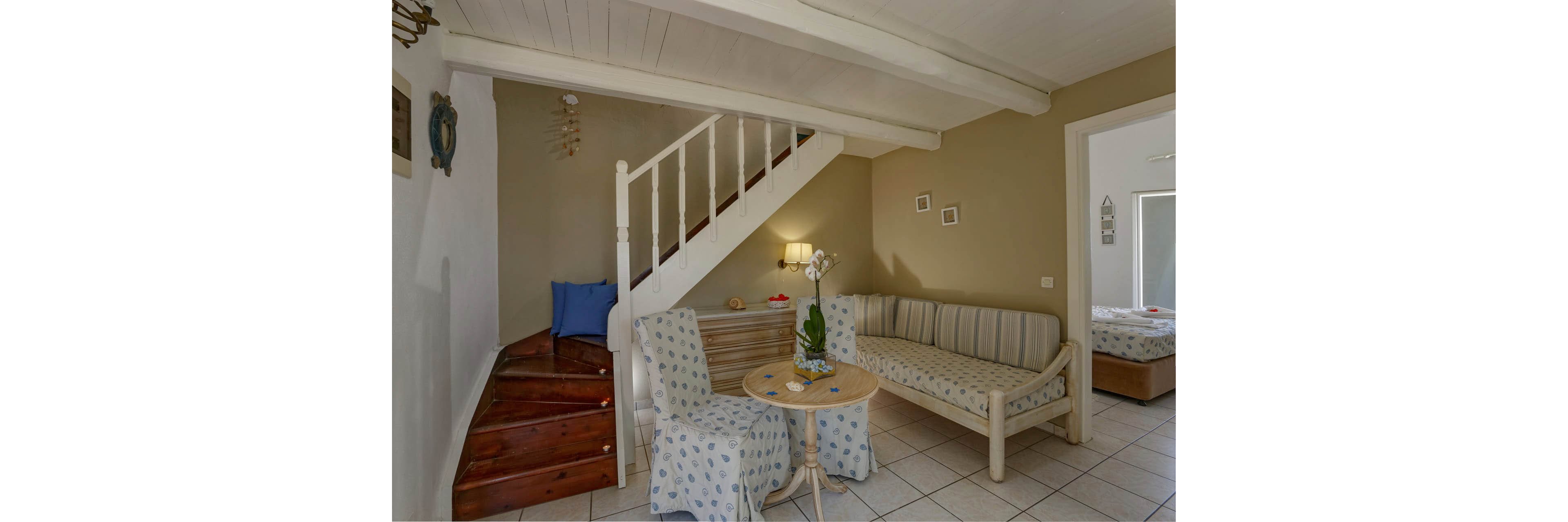 Small living room features a striped sofa and two slipcovered armchairs around a round wooden table. A white staircase with wooden steps leads to an upper floor, adding architectural interest to the space. Neutral beige walls and white ceiling beams create a calm, coastal-inspired atmosphere, accented by blue cushions and patterned fabrics. In the background, an adjoining bedroom with a double bed is visible through an open doorway.