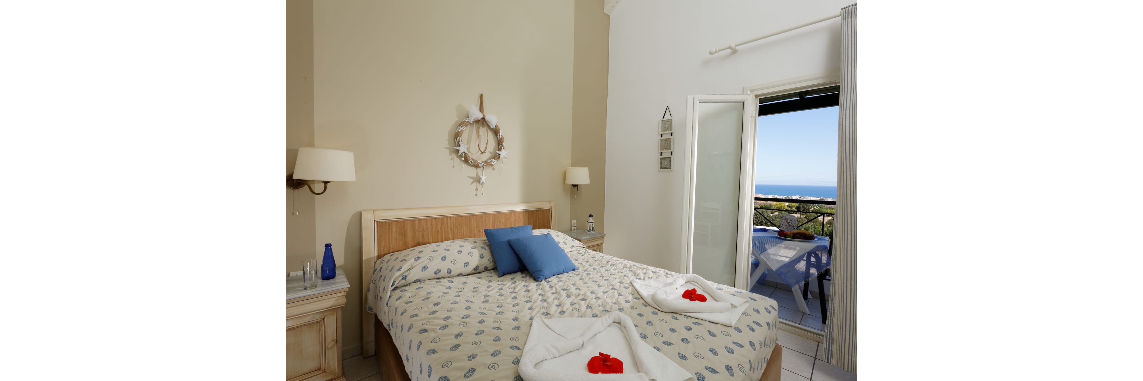 Bright bedroom interior features a large bed with patterned bedding and blue accent pillows. Light wooden furniture and neutral walls create a calm, airy atmosphere. Towels folded on the bed with red flower decorations add a welcoming touch. An open door leads to a balcony with table and chairs overlooking the ocean and landscape.