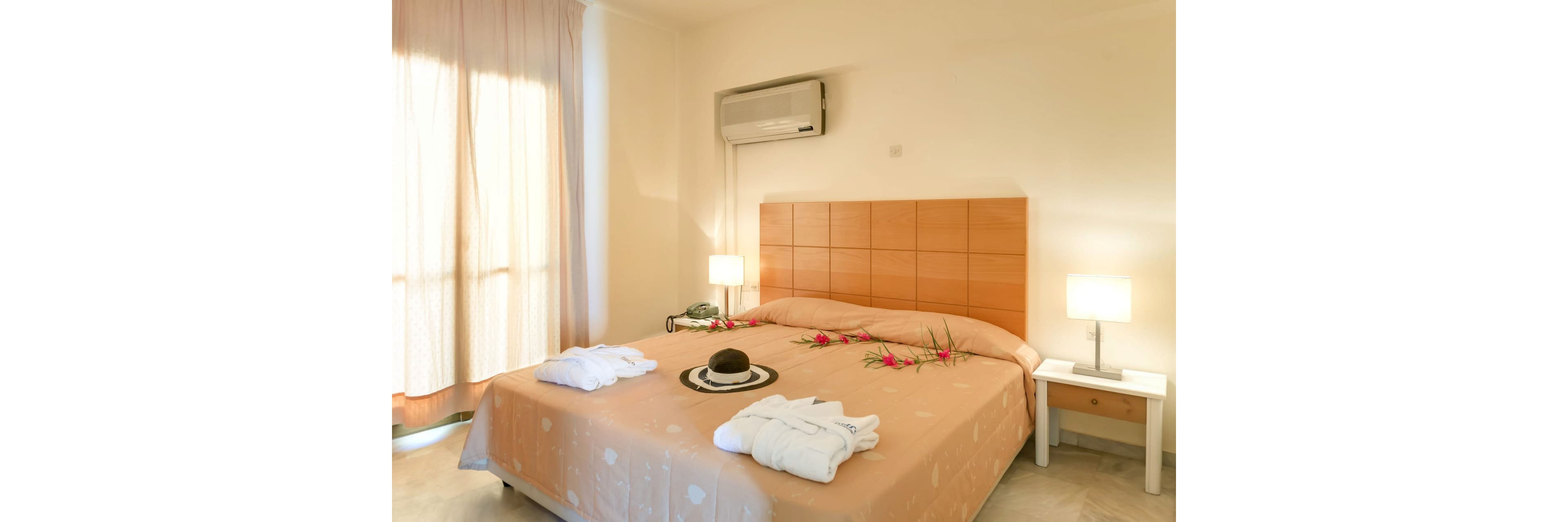 A neatly arranged double bed is the focal point of a bright hotel bedroom. The bedspread is beige and decorated with pink flowers and a hat placed on a tray. Two bedside tables with modern lamps flank the bed, adding warm ambient light. An air conditioning unit is mounted above the wooden headboard, and soft curtains filter daylight from the window.