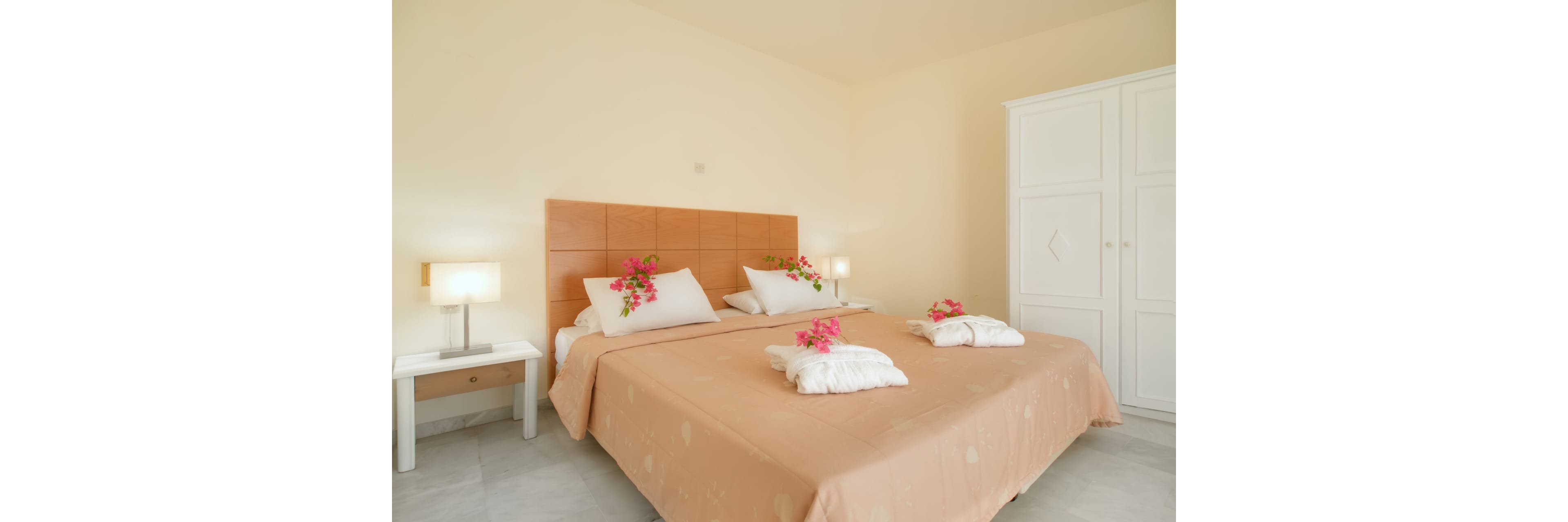 A neatly made double bed with a tan bedspread is the focal point of a bright, minimalist bedroom. White pillows and folded towels are decorated with vibrant pink flowers, adding a fresh, welcoming touch. Matching bedside tables with modern lamps flank the bed, while a large white wardrobe stands against the wall. The overall setting feels clean, airy, and inviting, suitable for a hotel or vacation rental.