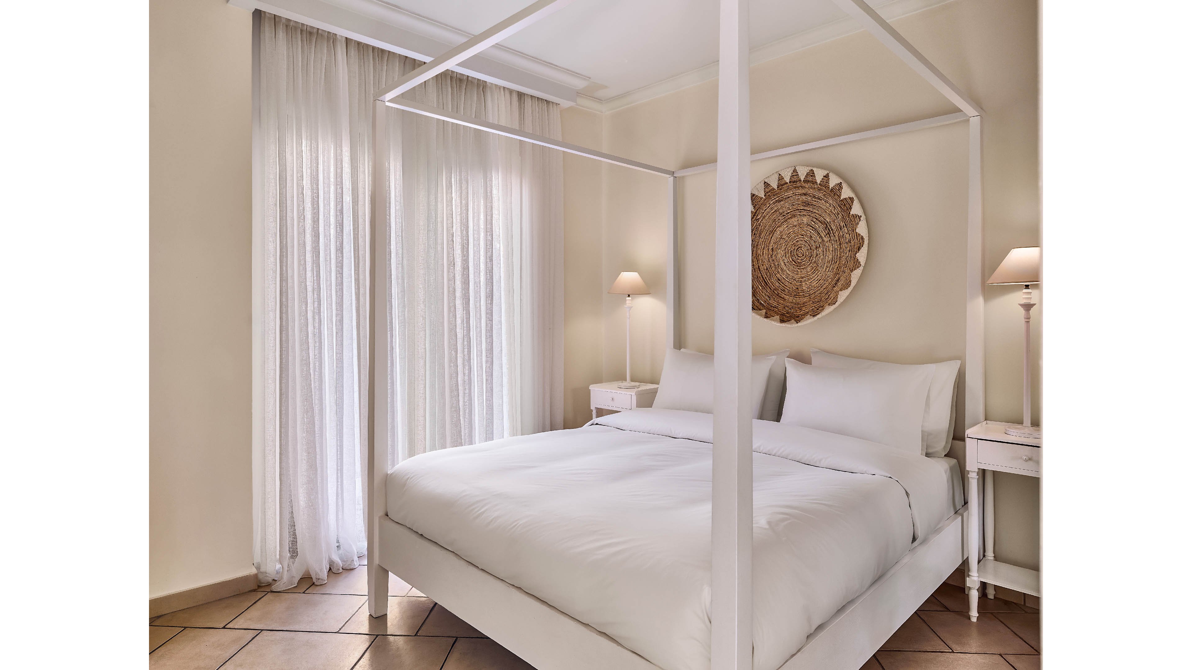 A neatly made white canopy bed is the focal point of this bright bedroom. Soft daylight filters through sheer curtains covering tall windows, creating a calm and airy atmosphere. Two small bedside tables with matching lamps flank the bed, while a woven circular wall decor adds texture above the headboard. Neutral tones and clean lines emphasize a minimalist, serene interior design.