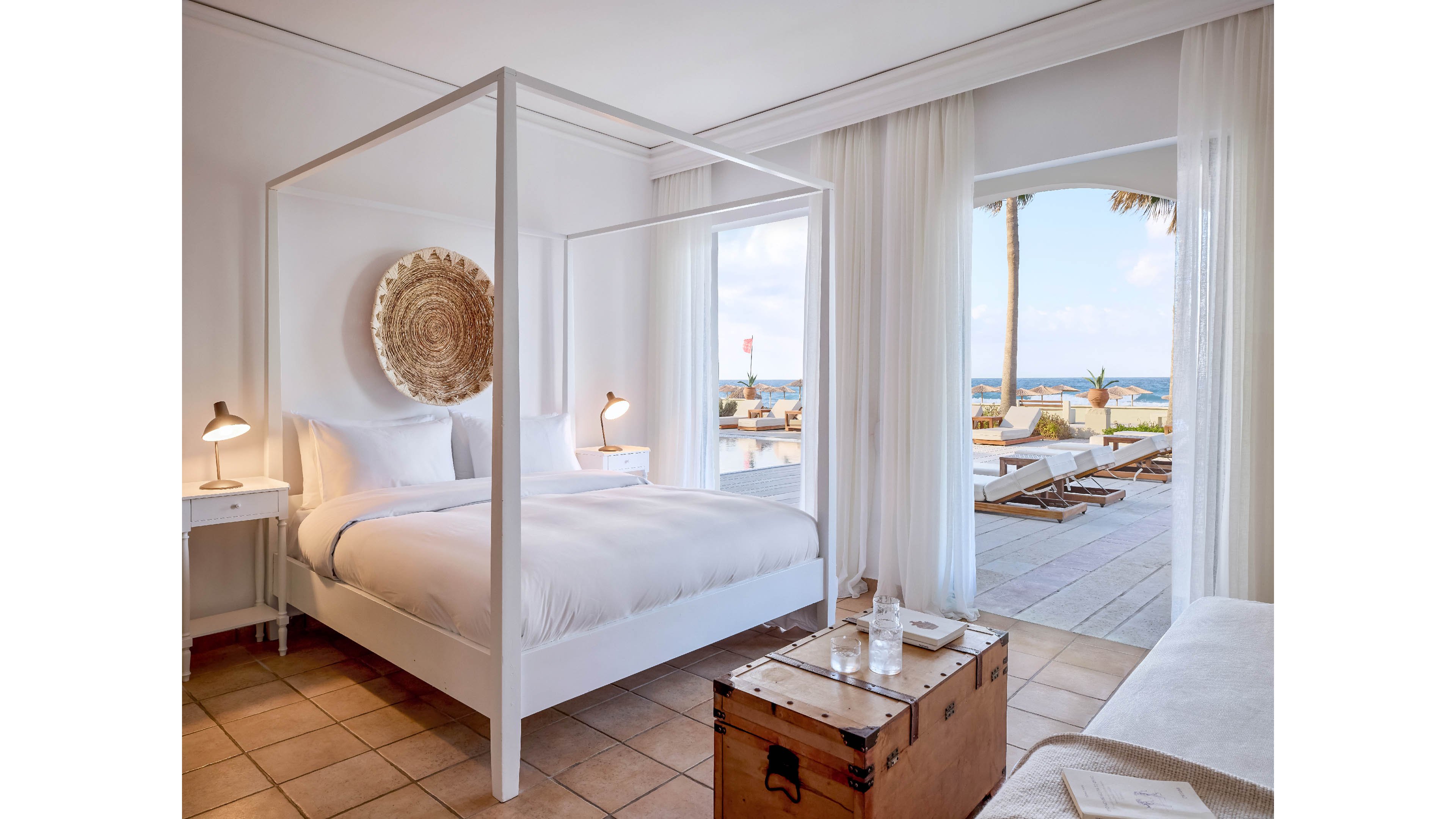 A bright, minimalist hotel bedroom features a white four-poster bed with crisp linens. Sheer curtains frame large open doors leading to a terrace with lounge chairs and a view of the sea. A rustic wooden trunk serves as a bedside table, holding a glass bottle and water glasses. Soft natural light and neutral tones create a calm, luxurious coastal atmosphere.