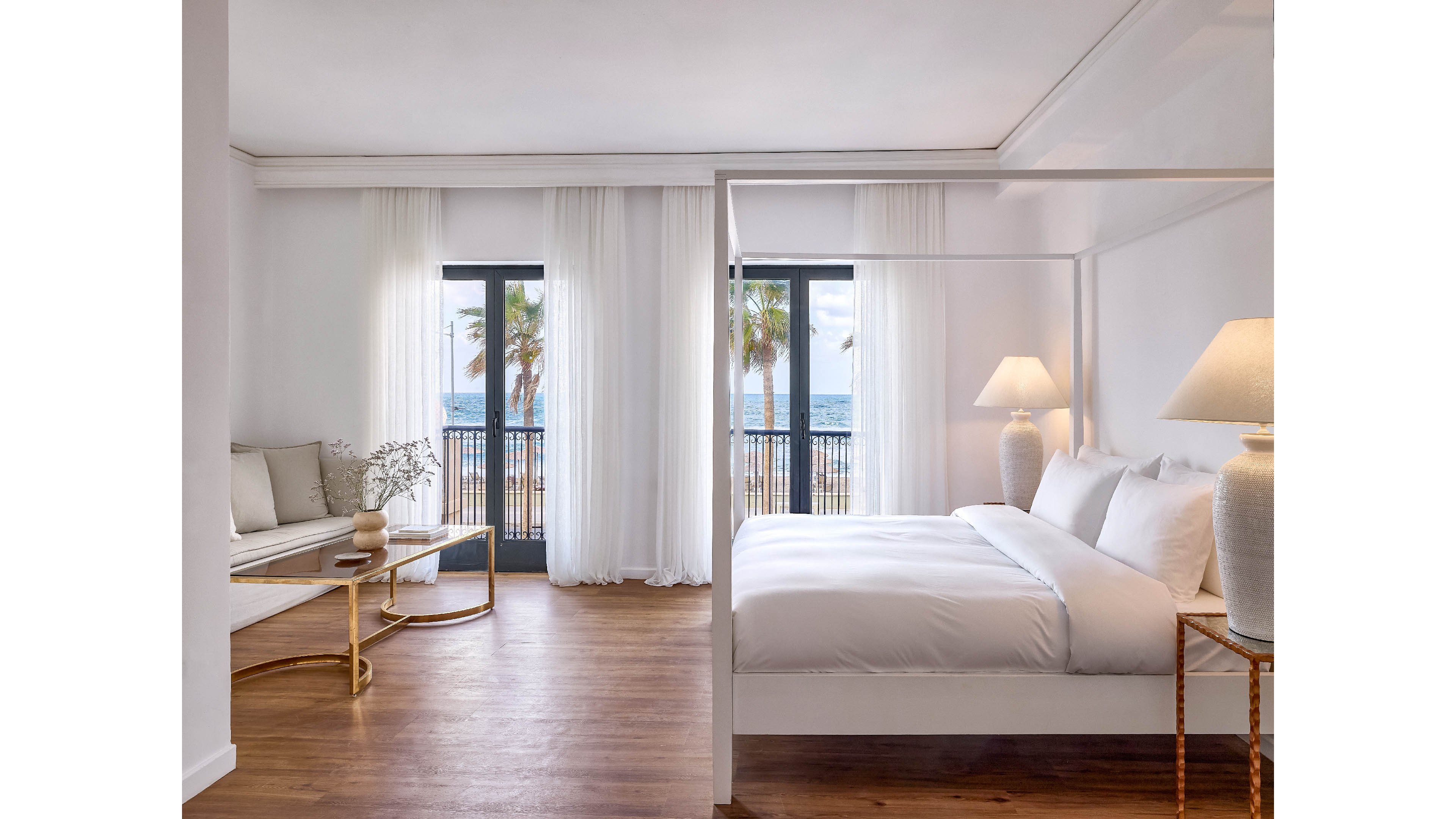 A spacious, minimalist hotel bedroom features a white four-poster bed and soft neutral furnishings. Large windows with sheer curtains open to a balcony overlooking palm trees and the ocean. Warm wooden floors contrast with the crisp white walls and bedding. Elegant table lamps and a small seating area create a serene, luxurious retreat.