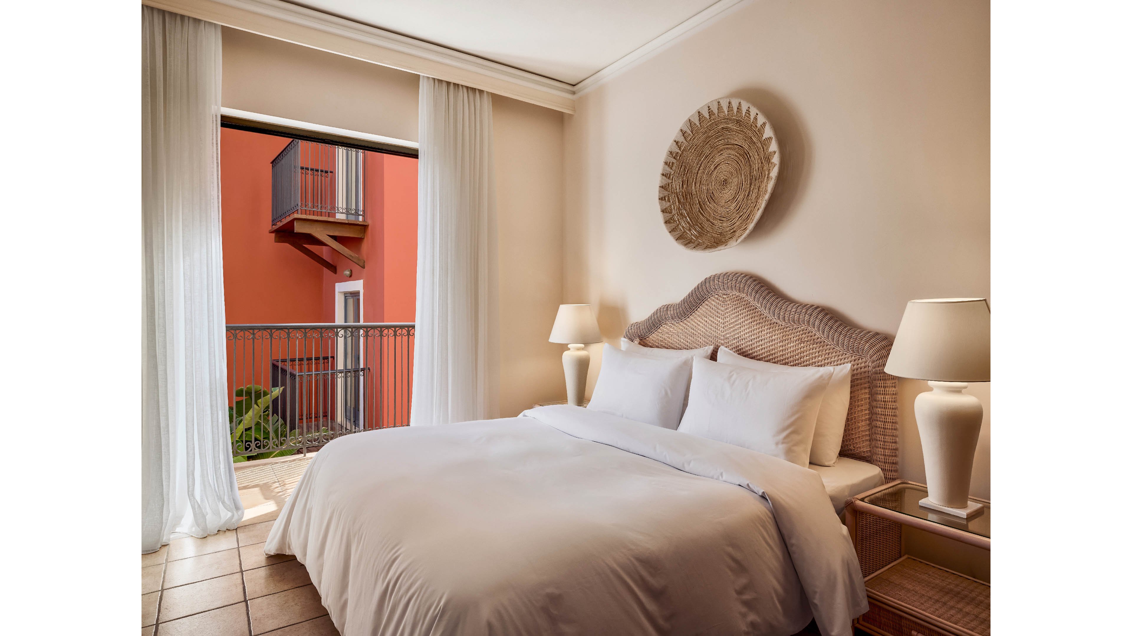 A neatly made double bed with crisp white linens is the focal point of this cozy hotel bedroom. The room features warm beige walls, wicker-style furniture, and matching bedside lamps. Sheer white curtains frame a balcony doorway that opens to an exterior view of a red building and railings. Soft natural light enhances the calm, inviting atmosphere of the interior space.