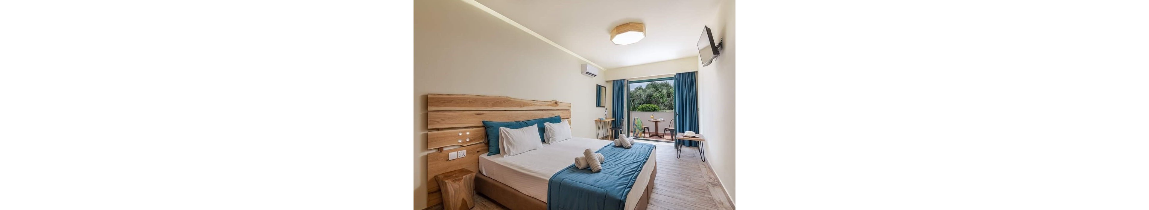 A contemporary hotel room features a large double bed with white linens and blue accents. Natural wood furniture and flooring create a warm, minimalist aesthetic. The space includes a wall-mounted television, air conditioner, and a small desk area. Glass doors open to a balcony with outdoor seating and lush greenery visible outside.