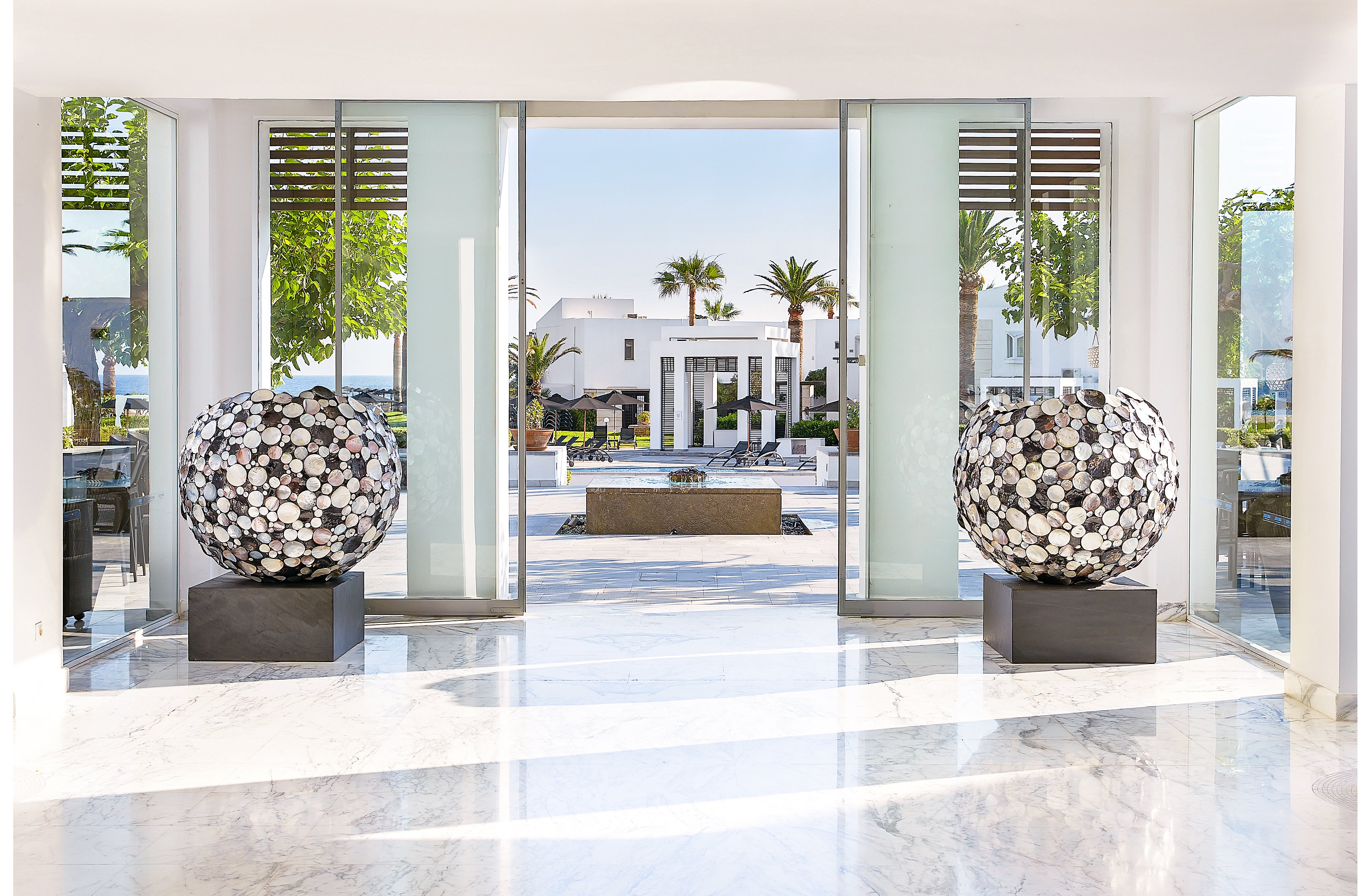 A bright and spacious lobby featuring two large decorative spheres on black stands. The setting opens to an outdoor area with palm trees, a fountain, and white architectural structures. The polished marble flooring and glass doors enhance the modern and luxurious ambiance.