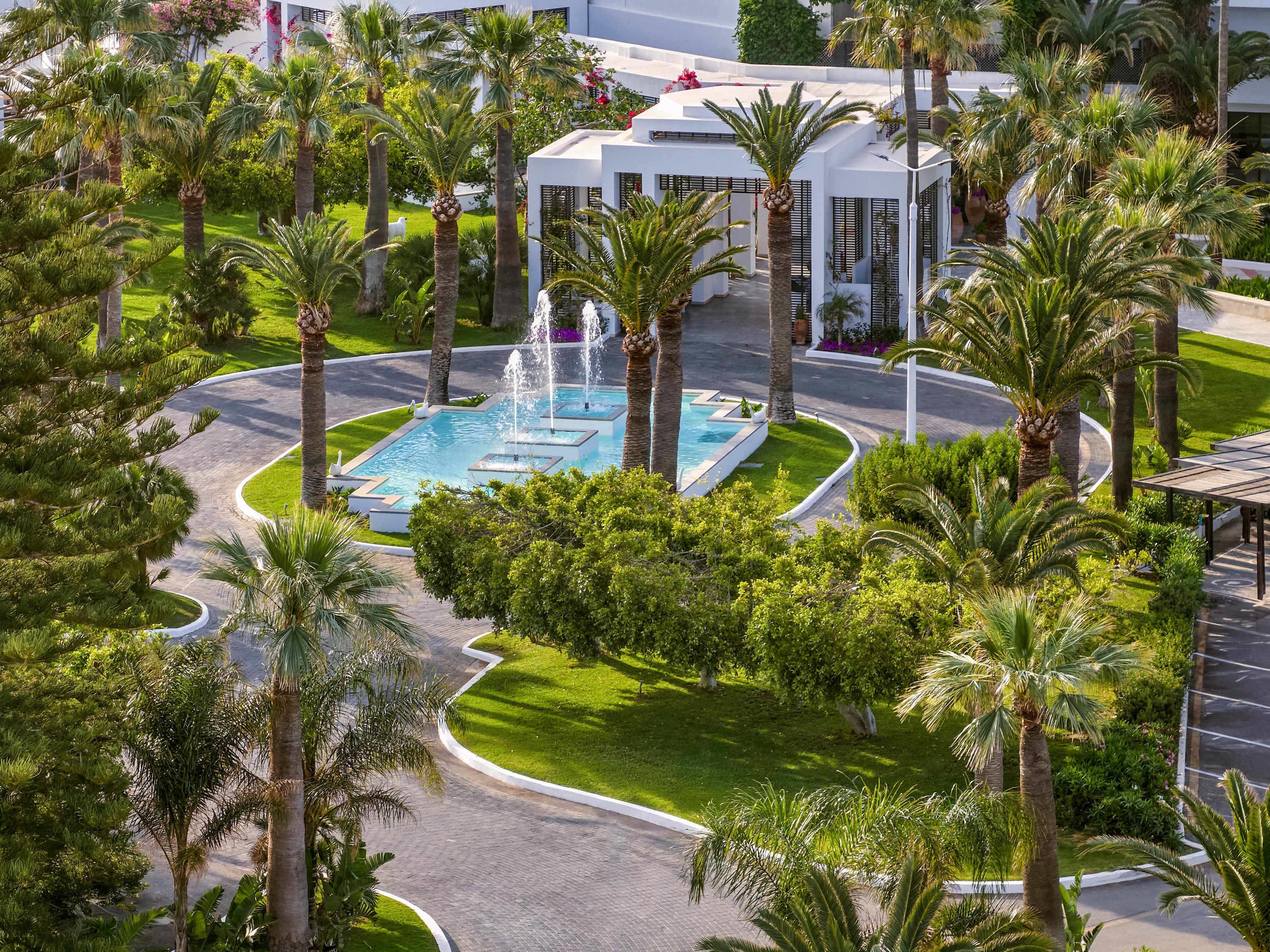A lush resort entrance features a circular fountain surrounded by tall palm trees and manicured greenery. Curving driveways lead vehicles around the central water feature toward a modern white building with large glass windows. The scene is bathed in bright daylight, highlighting the vibrant green lawns and clear blue water in the fountain. The overall atmosphere feels luxurious, tranquil, and welcoming.