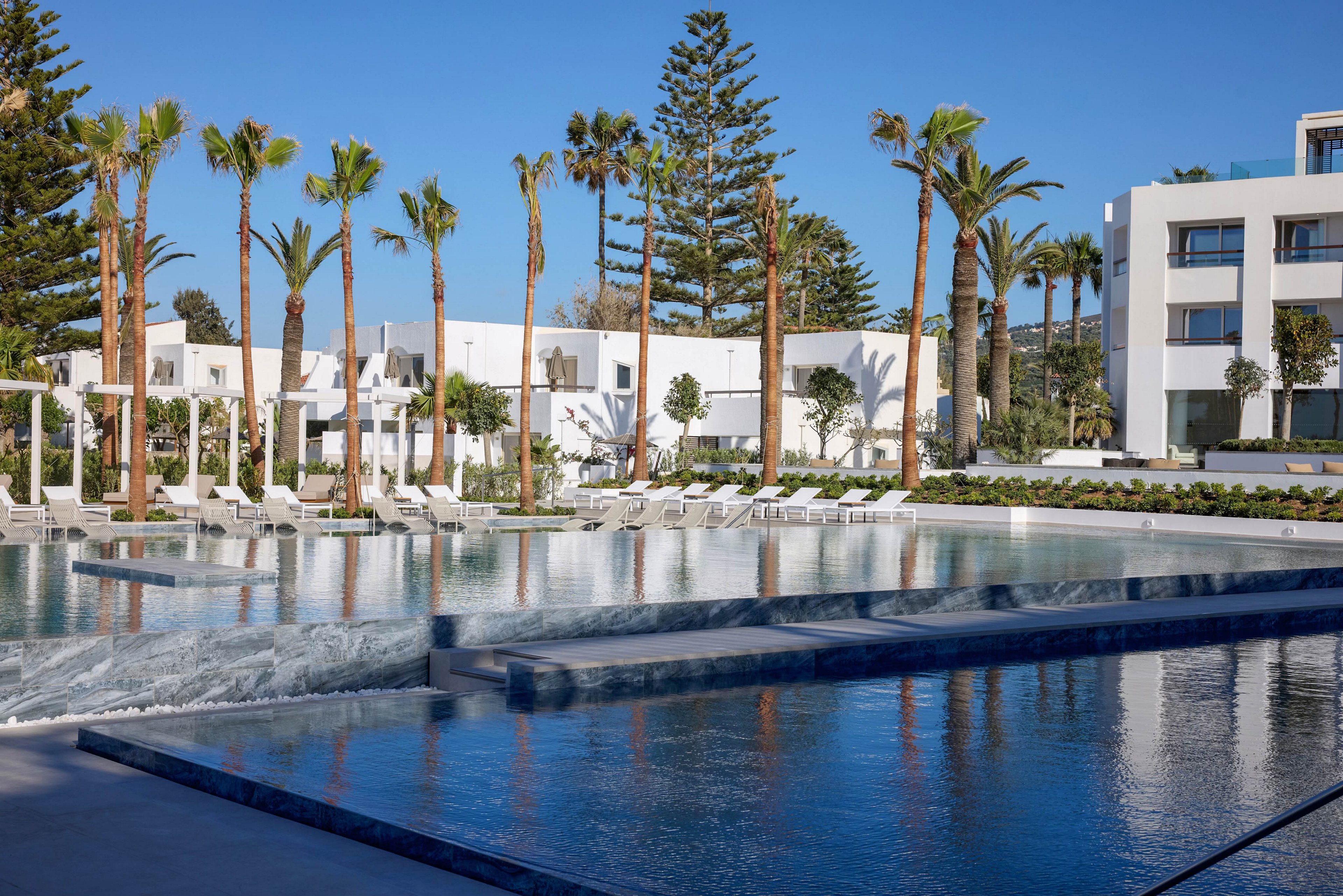A contemporary resort features a large, tranquil swimming pool surrounded by white sun loungers and umbrellas. Tall palm trees and manicured greenery line the poolside, adding a tropical feel to the modern white buildings in the background. The clear blue water reflects the architecture and sky, creating a serene vacation atmosphere. The scene is bright and sunny, suggesting a warm, relaxing holiday destination.