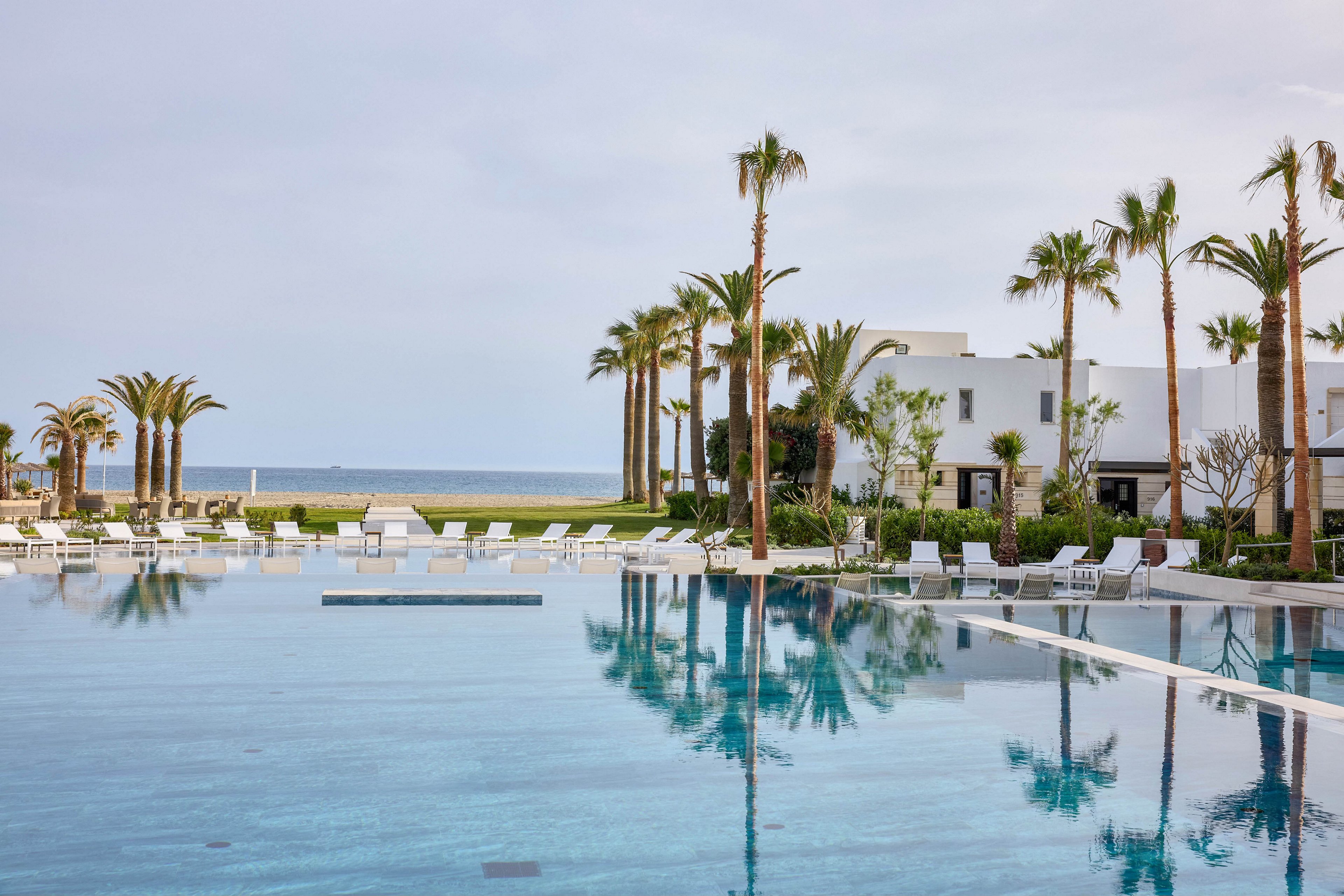 A large, tranquil swimming pool reflects tall palm trees and modern white resort buildings. Numerous white lounge chairs and umbrellas line the poolside, suggesting a relaxing vacation setting. The ocean is visible in the background under a soft, overcast sky. The overall scene feels calm, minimal, and luxurious, emphasizing leisure by the sea.