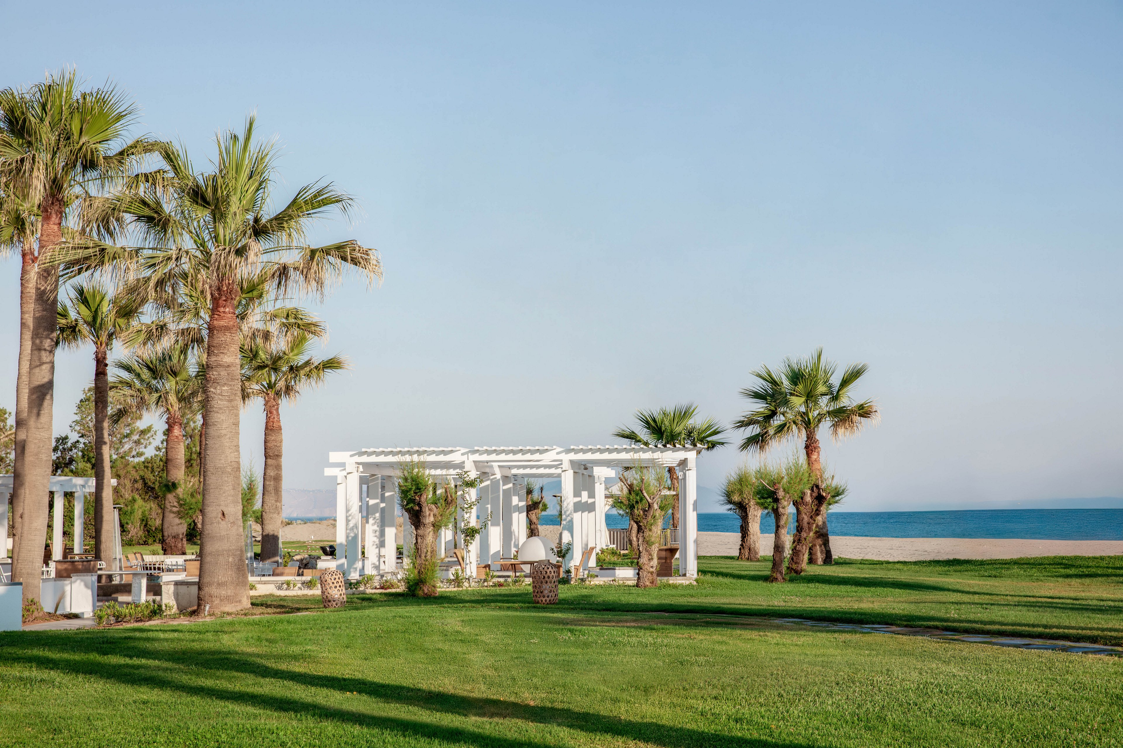 A white pergola stands on a manicured green lawn surrounded by tall palm trees. The scene overlooks a calm blue sea under a clear sky. Outdoor seating and decorative elements suggest a relaxing coastal retreat or event space. The overall atmosphere is bright, open, and tranquil.