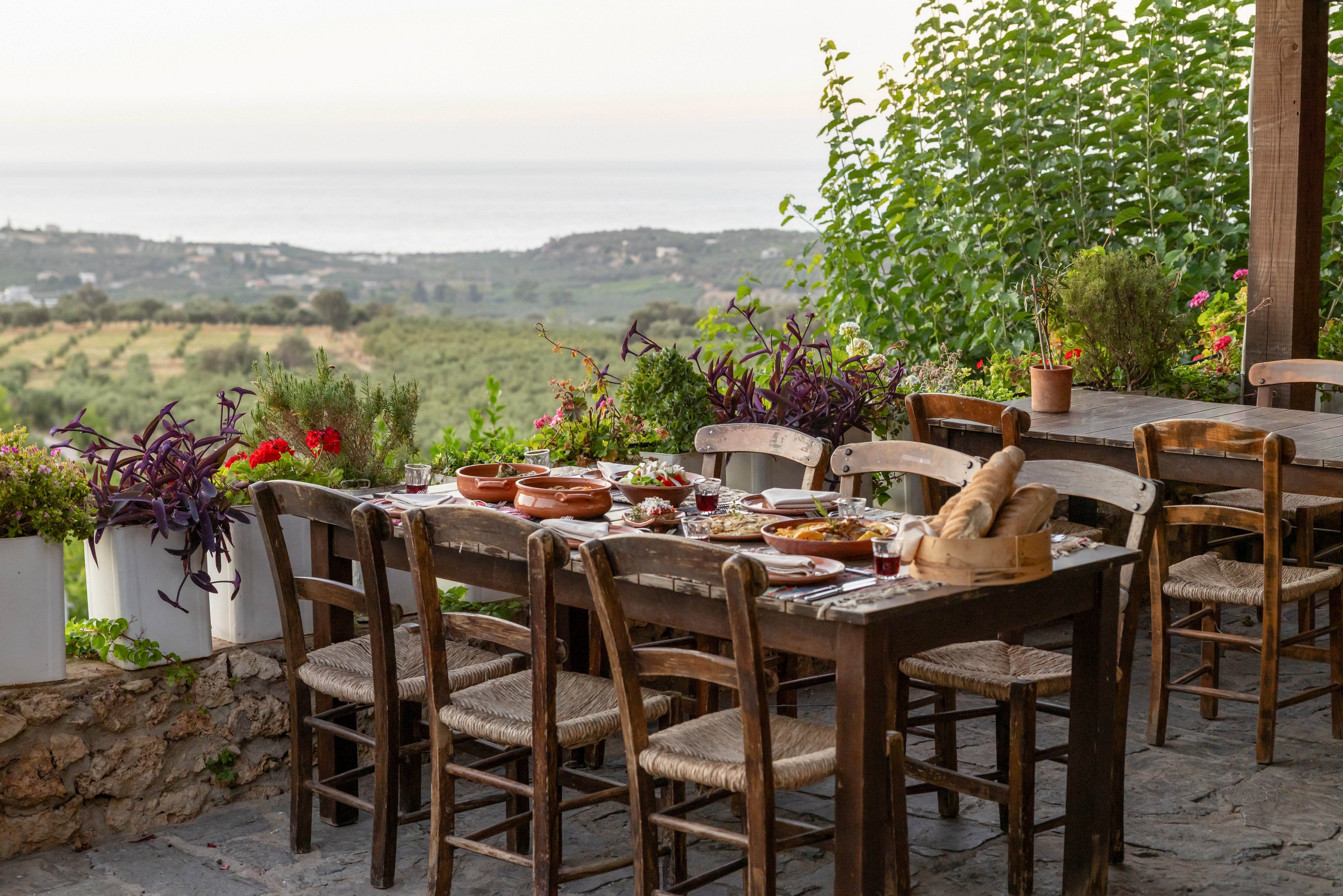 A long wooden dining table is set outdoors with plates, glasses, and rustic serving dishes ready for a shared meal. Wooden chairs surround the table on a stone patio, suggesting a relaxed gathering. Lush potted plants frame the scene, and rolling countryside stretches into the distance under a soft sky. The overall atmosphere is warm, inviting, and tranquil, ideal for an al fresco feast.