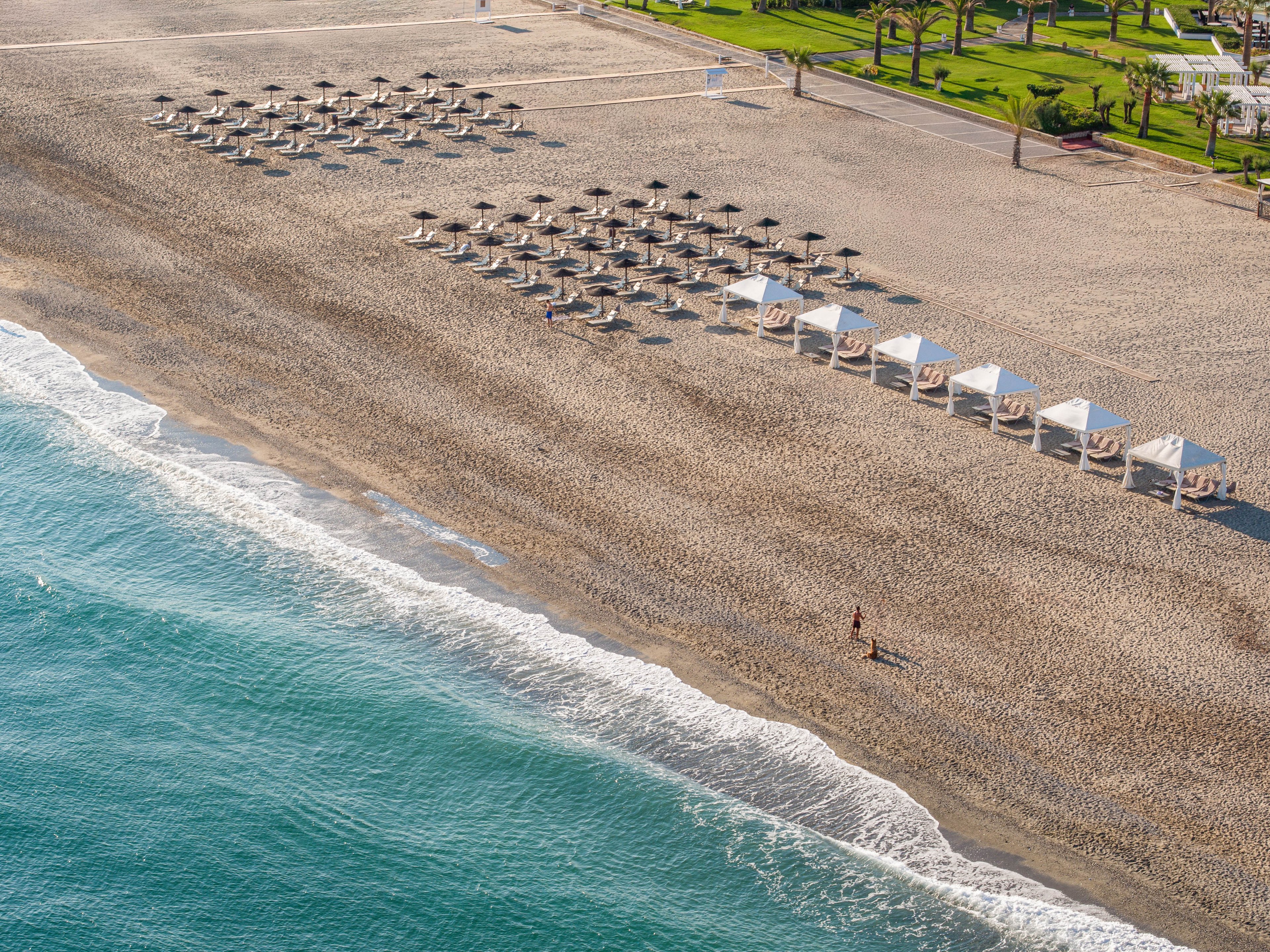 A wide sandy beach stretches along a calm turquoise sea, lined with neat rows of white sun umbrellas and cabanas. The resort area appears quiet and mostly empty, with only a single person walking near the shoreline. Gentle waves roll onto the sand, creating a soft white surf edge. Green landscaped grounds and pathways are visible in the distance beyond the beach.