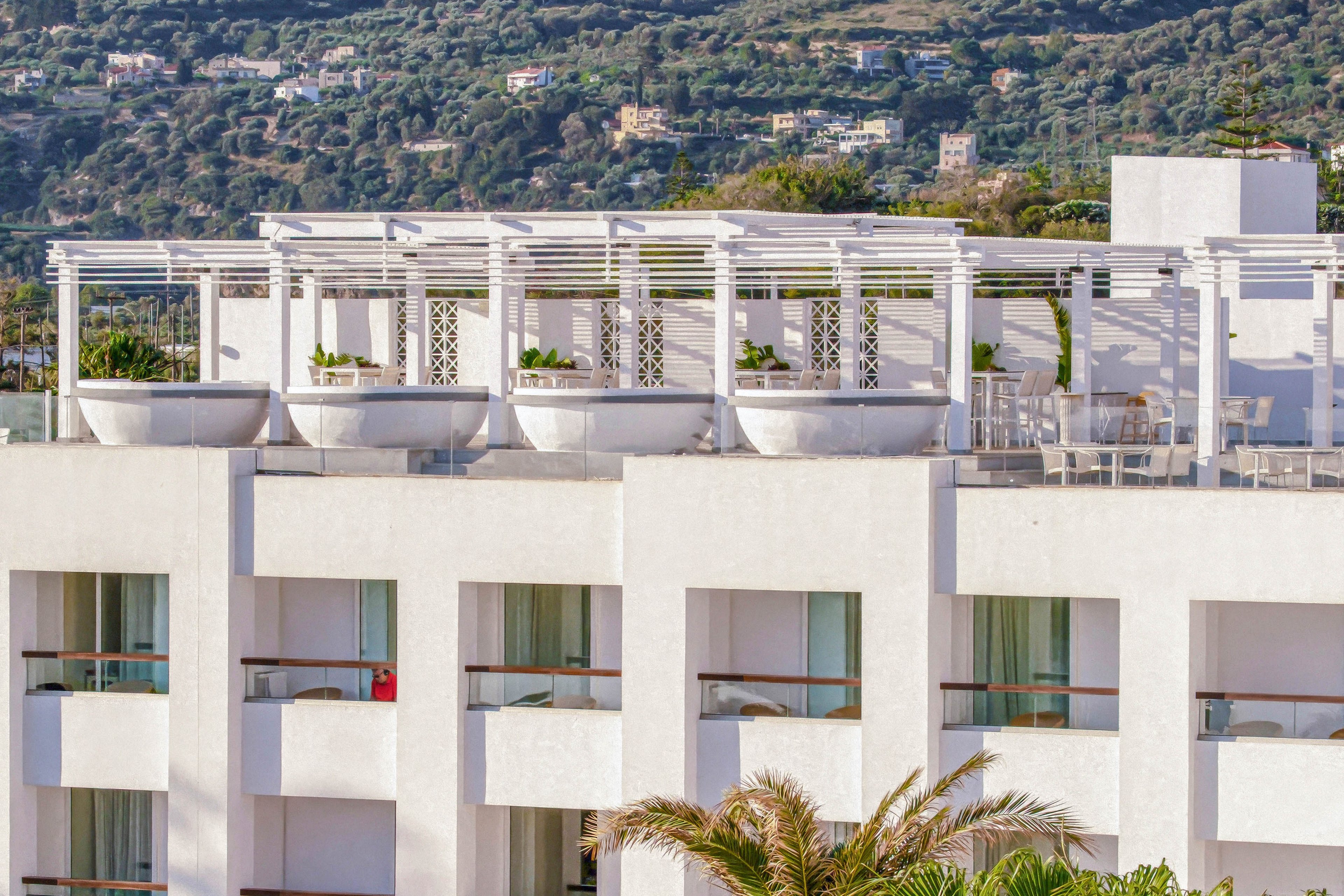 A contemporary white resort building stands prominently in a lush coastal setting. The structure features multiple floors with balconies and a rooftop terrace framed by clean geometric lines. Palm trees and greenery surround the hotel, with outdoor seating and umbrellas visible at ground level. Rolling green hills and mountains rise in the background under a partly cloudy sky, creating a relaxed vacation atmosphere.