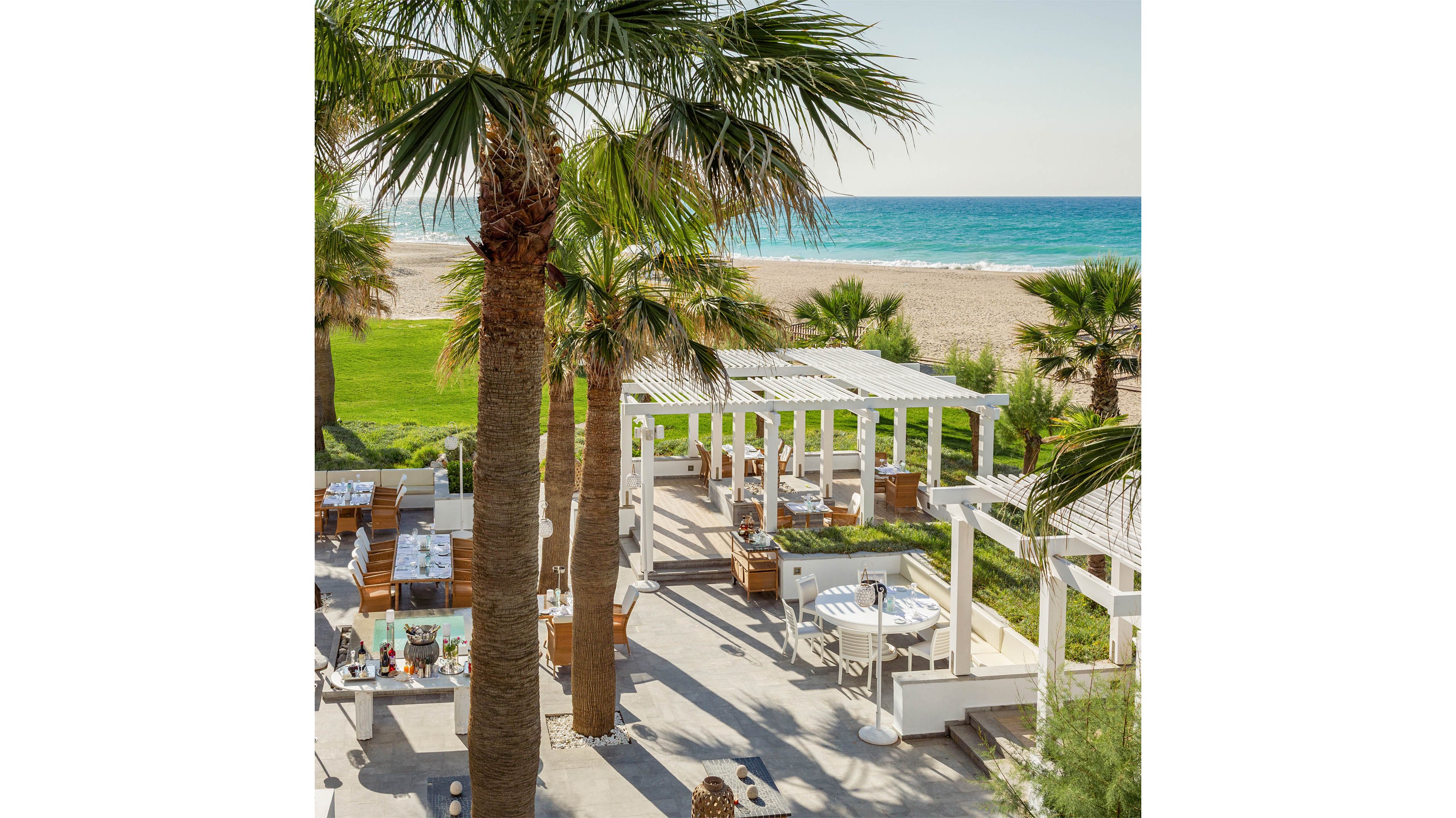 An open-air restaurant is set among tall palm trees overlooking a turquoise sea. White pergolas shade neatly arranged wooden tables and chairs on a stone terrace. The setting includes manicured green lawns leading to a sandy beach in the background. Bright daylight and clear skies create a relaxed, upscale coastal dining atmosphere.