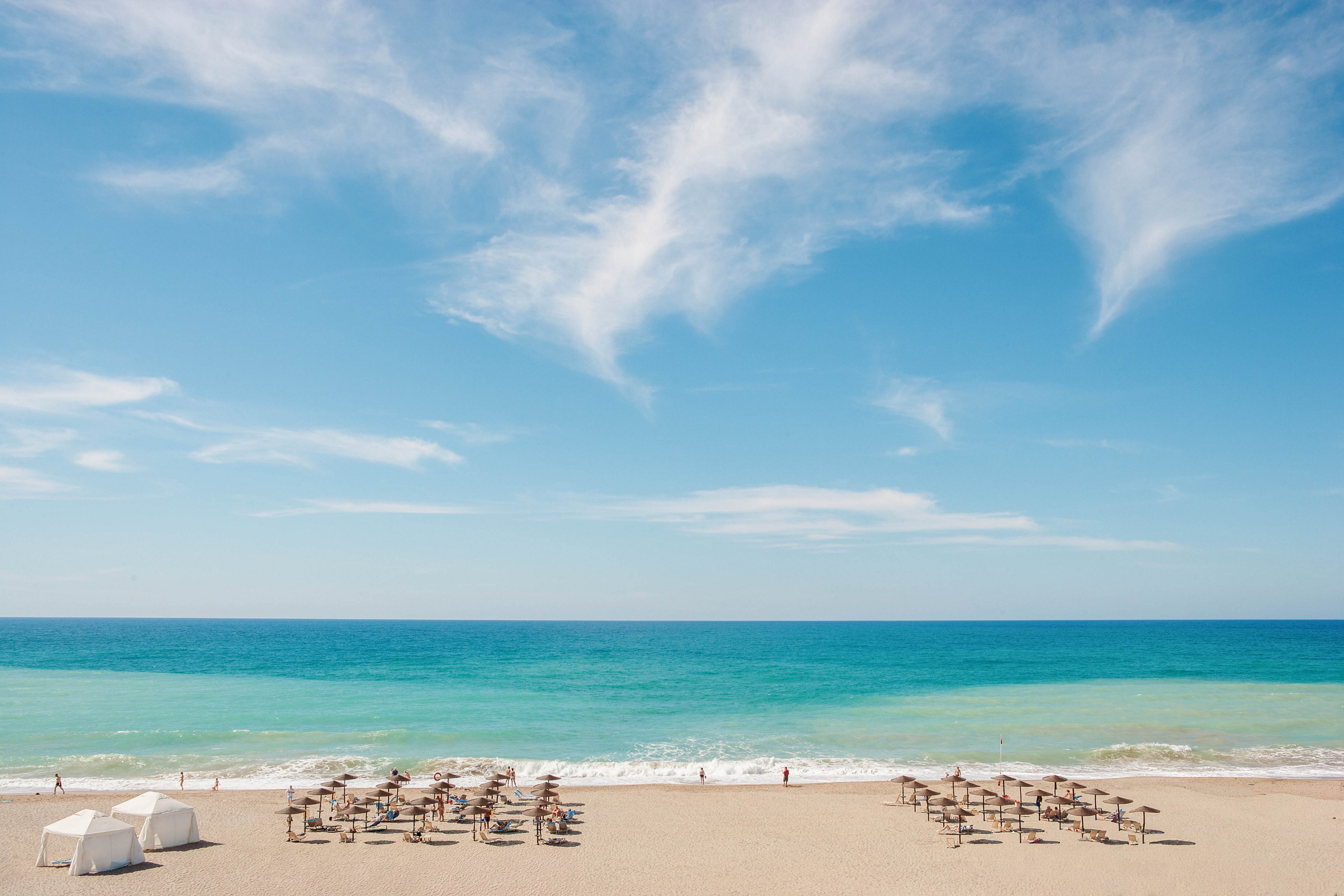 A wide sandy beach stretches out toward a calm turquoise sea under a bright blue sky with wispy clouds. Neatly arranged rows of sun umbrellas and loungers line the shore, suggesting a resort or holiday destination. A few small figures walk along the waterline, adding a sense of scale and leisure. Two white cabana-style tents sit on the sand in the foreground, enhancing the relaxed, summery atmosphere.