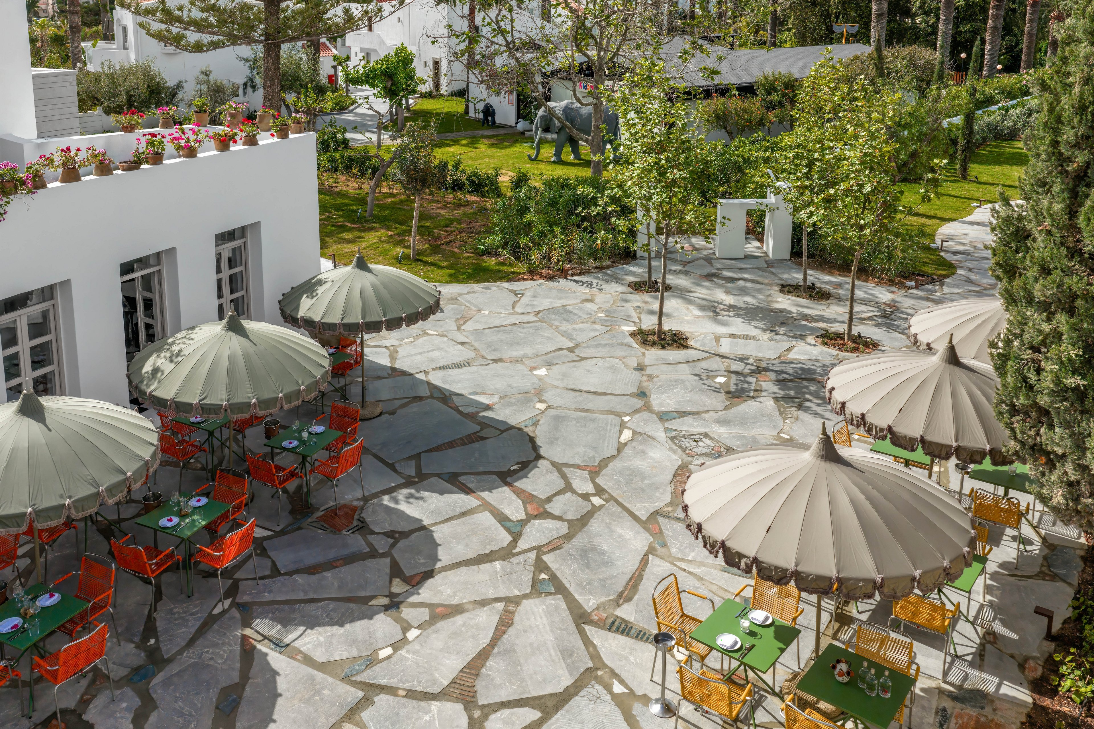 An outdoor café terrace is set in a stone-paved courtyard beside a white building. Several tables with green tablecloths and orange chairs sit under large beige umbrellas. The area is surrounded by lush greenery, trees, and manicured lawns, creating a tranquil garden atmosphere. In the background, decorative elements and pathways add depth to the resort-like setting.