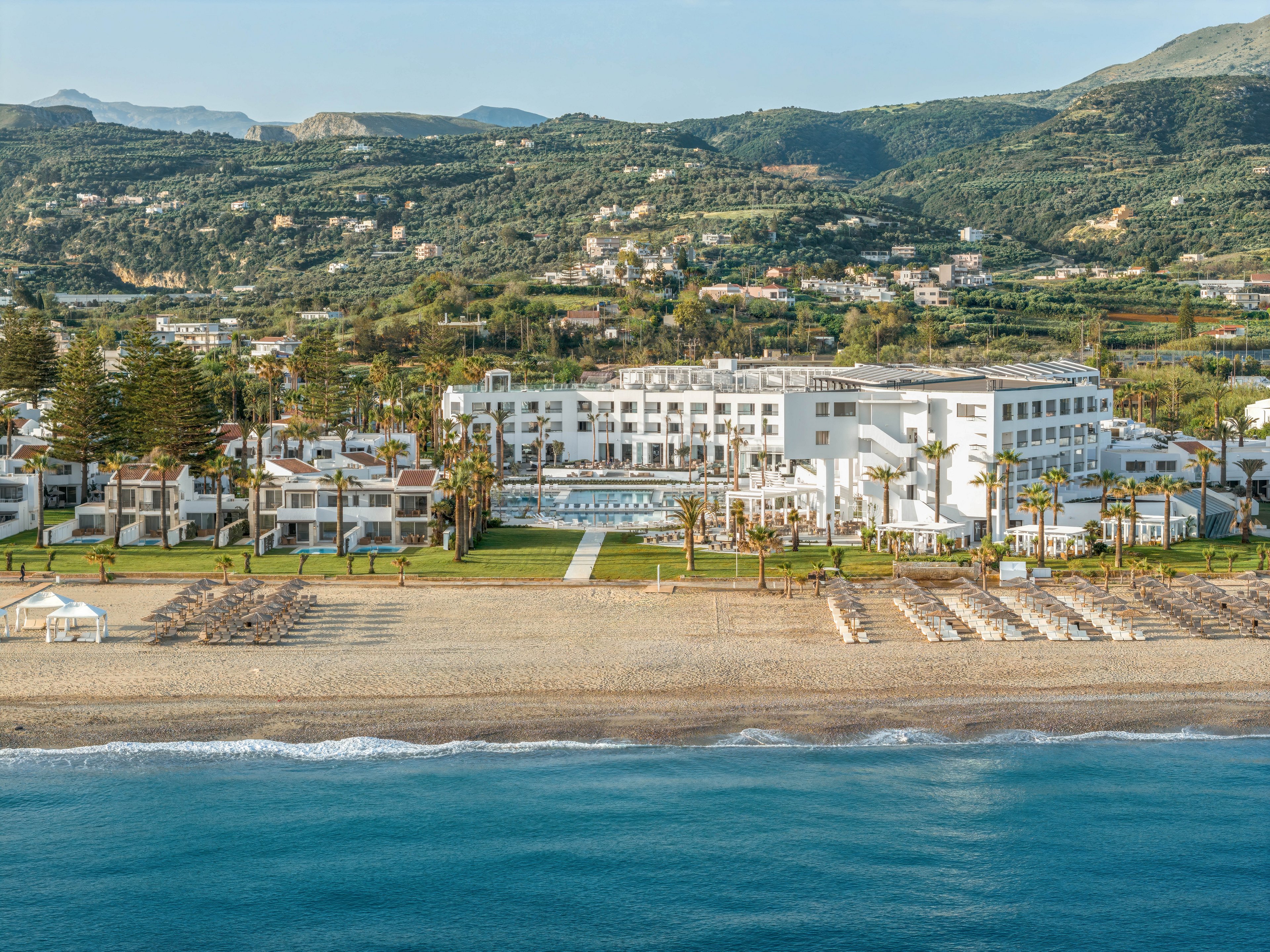 A large white beachfront resort stretches along a sandy shore facing calm blue water. Rows of sun loungers and umbrellas line the beach in front of manicured lawns and palm trees. The resort buildings surround a central pool area and are backed by lush green hills dotted with houses. The scene suggests a tranquil holiday destination with a mix of seaside and mountain scenery.