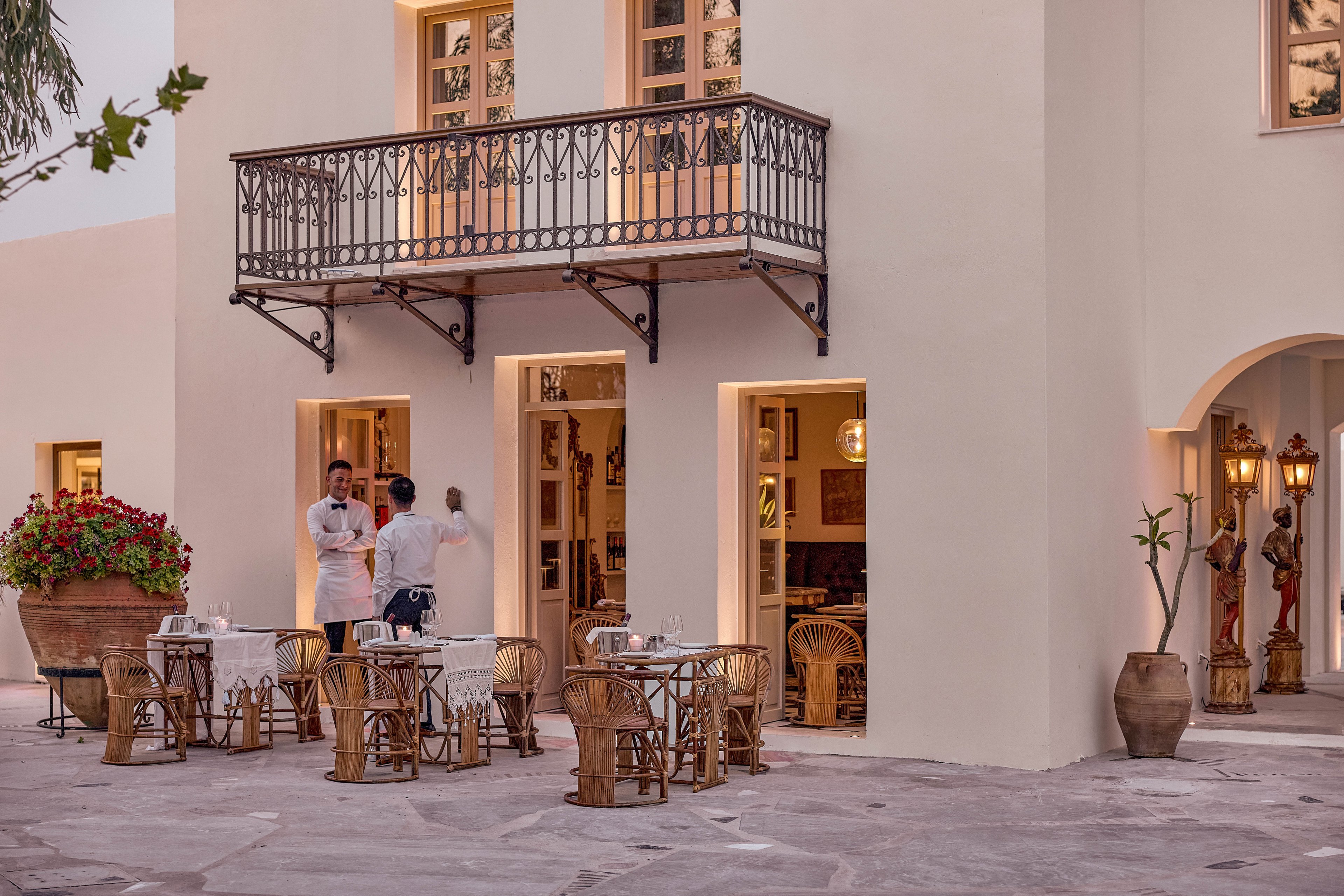 An elegant outdoor restaurant terrace is set in front of a light-colored Mediterranean-style building. Two staff members in white uniforms stand near the entrance, preparing for guests. Wicker chairs and neatly arranged tables with white tablecloths create an inviting dining area. Warm lighting from windows and lanterns, potted plants, and decorative statues enhance the relaxed evening ambiance.