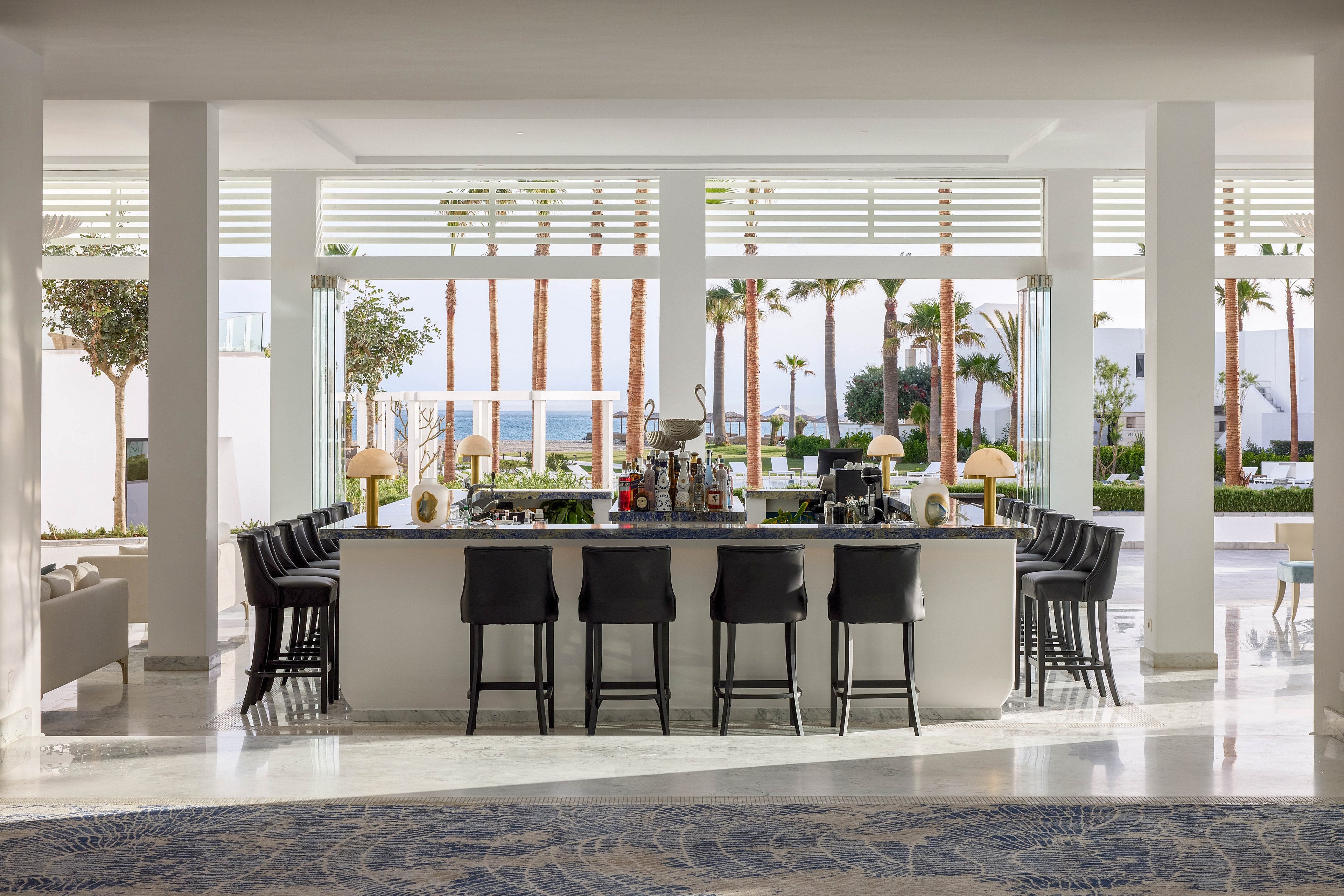 A sleek, modern bar with black stools stands at the center of a bright resort lounge. Large windows and open slats reveal tall palm trees and a distant view of the sea. Neutral white walls and polished floors create an airy, minimalist atmosphere. Decorative lamps and bar accessories add subtle detail to the sophisticated coastal setting.