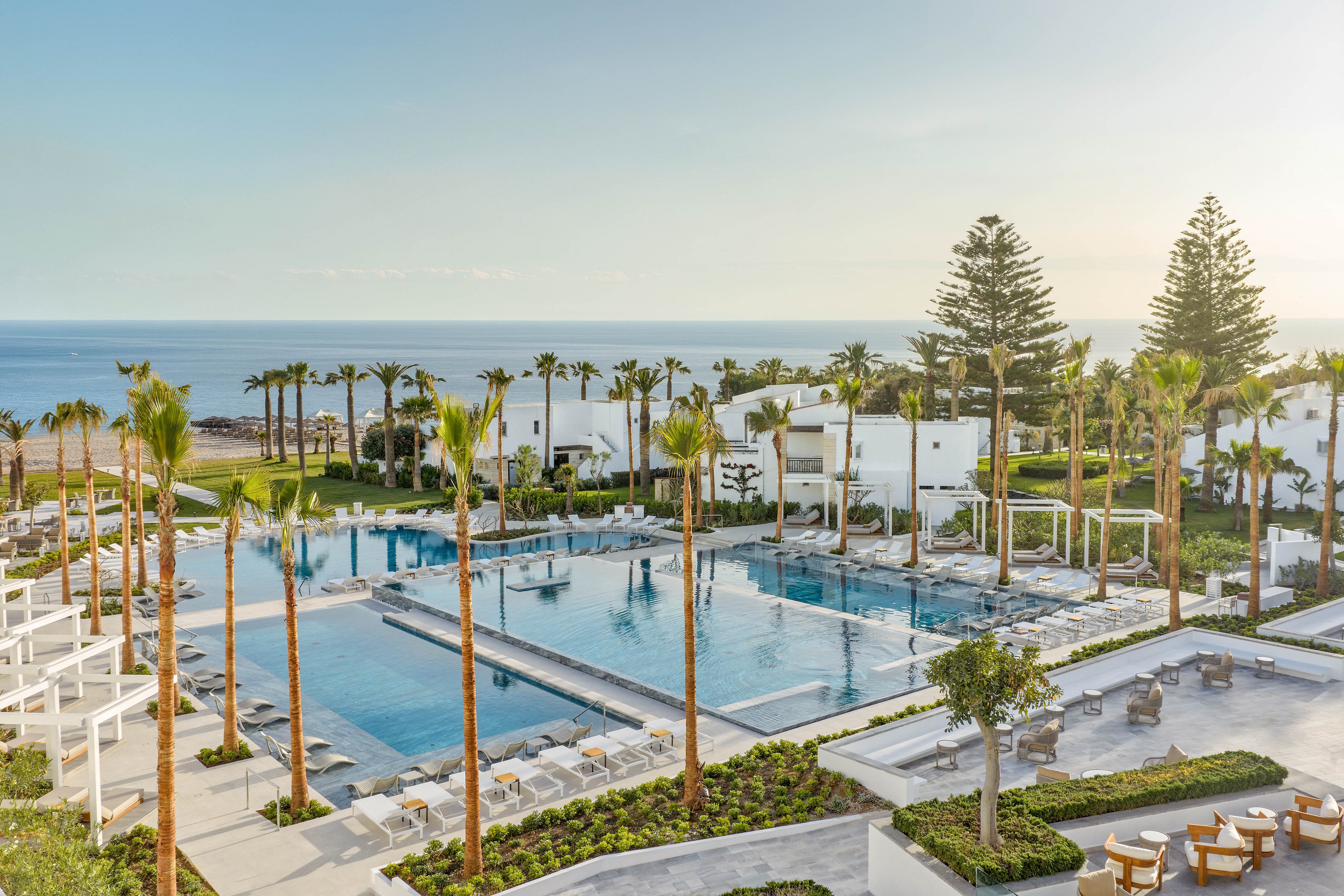 A modern seaside resort features expansive swimming pools surrounded by tall palm trees and white contemporary buildings. Sun loungers and umbrellas line the pool edges, creating a relaxing vacation atmosphere. The scene overlooks a calm ocean horizon under a clear sky. Outdoor seating areas and manicured gardens enhance the upscale, tranquil setting.