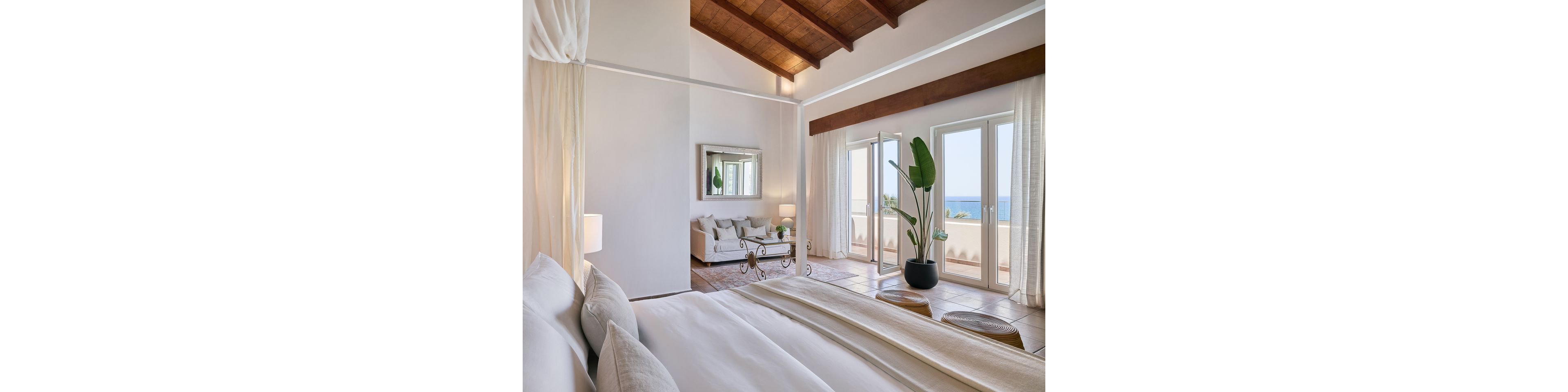 A spacious, sunlit bedroom suite features a large bed in the foreground and a cozy sitting area with a sofa and glass coffee table in the background. Floor-to-ceiling windows and glass doors open to a balcony with a clear view of the ocean. The room has high wooden ceilings, light neutral walls, and tiled floors that enhance the airy, coastal feel. Sheer curtains, indoor plants, and soft textiles create a serene, relaxing atmosphere.