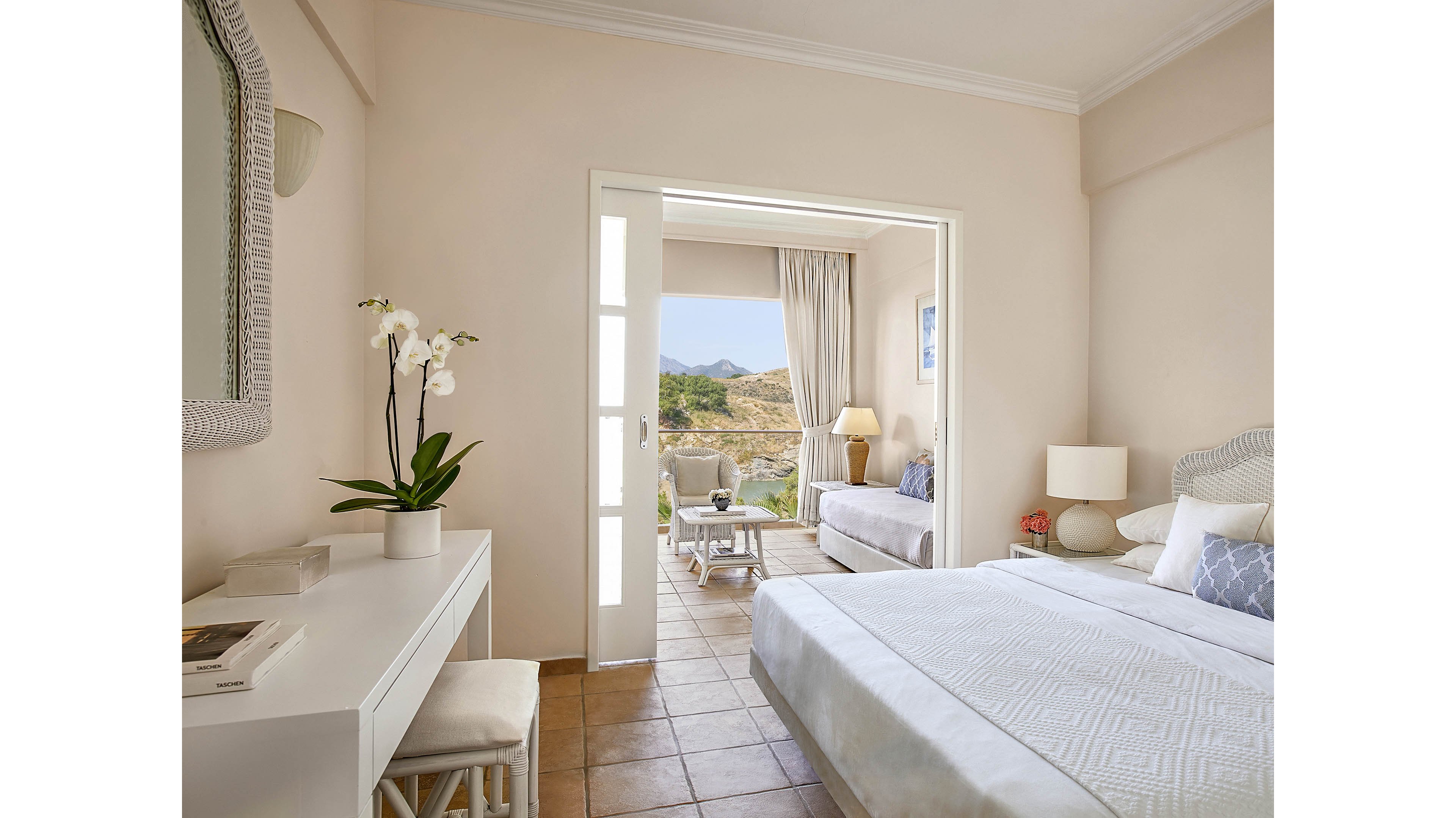 A spacious, sunlit hotel room features a neatly made bed, a white desk with an orchid plant, and a cozy seating area. The room opens to a terrace with outdoor furniture, overlooking scenic mountains and greenery. Soft neutral tones and minimalist decor create a calm, inviting atmosphere. No visible people or text are present in the image.