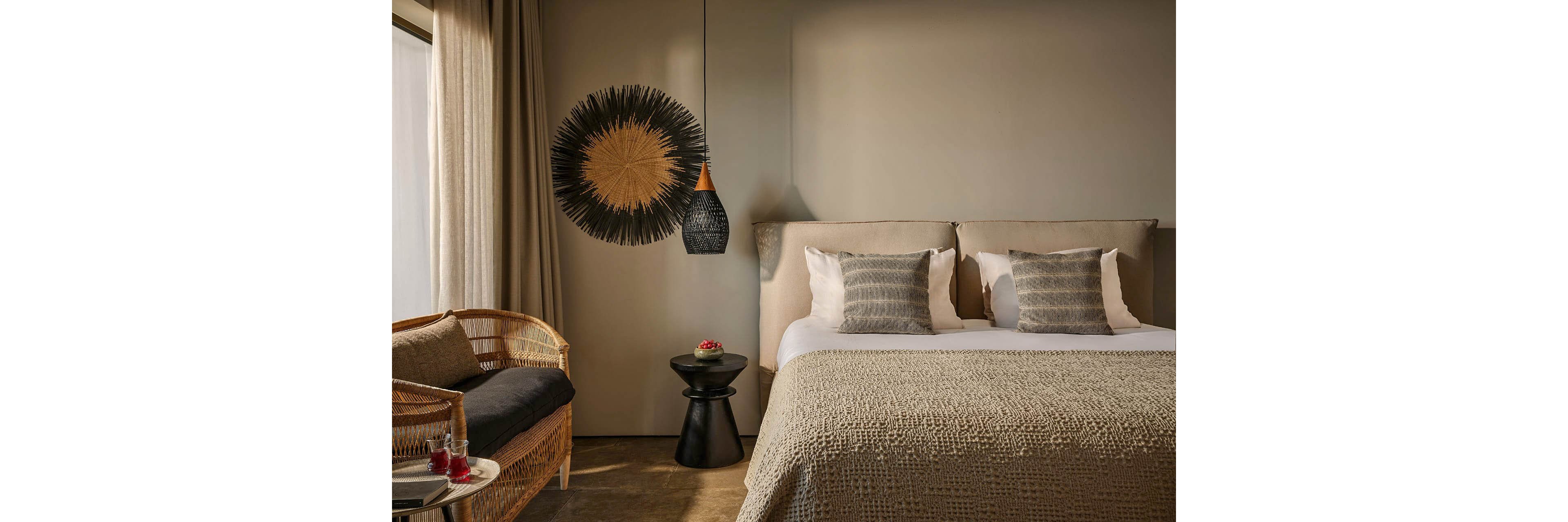 A contemporary bedroom features a neatly made double bed with textured beige bedding and matching pillows. The room includes a woven rattan daybed by the window with sheer curtains allowing soft natural light. A black side table with a small red floral arrangement sits between the bed and a sculptural wall hanging and pendant light. The overall design emphasizes warm neutral tones, natural materials, and minimalist styling for a calm, inviting atmosphere.