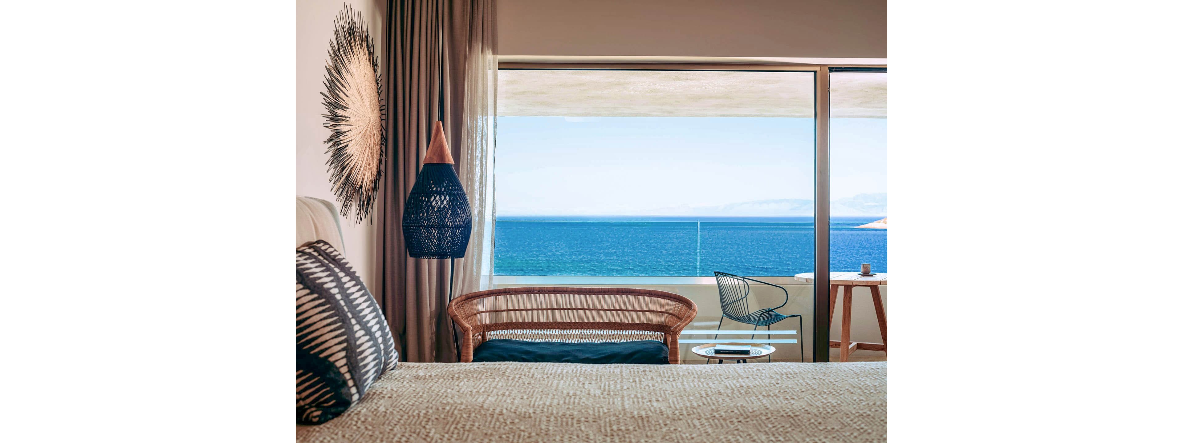 A contemporary hotel room features a bed with patterned pillows and textured bedding in the foreground. Natural materials like a woven headboard, wicker bench, and hanging lamp add warmth to the interior. Large floor-to-ceiling windows open to a balcony with minimalist chairs and a small table overlooking a calm blue sea. Soft neutral tones inside contrast with the vivid ocean and sky outside, creating a serene vacation atmosphere.