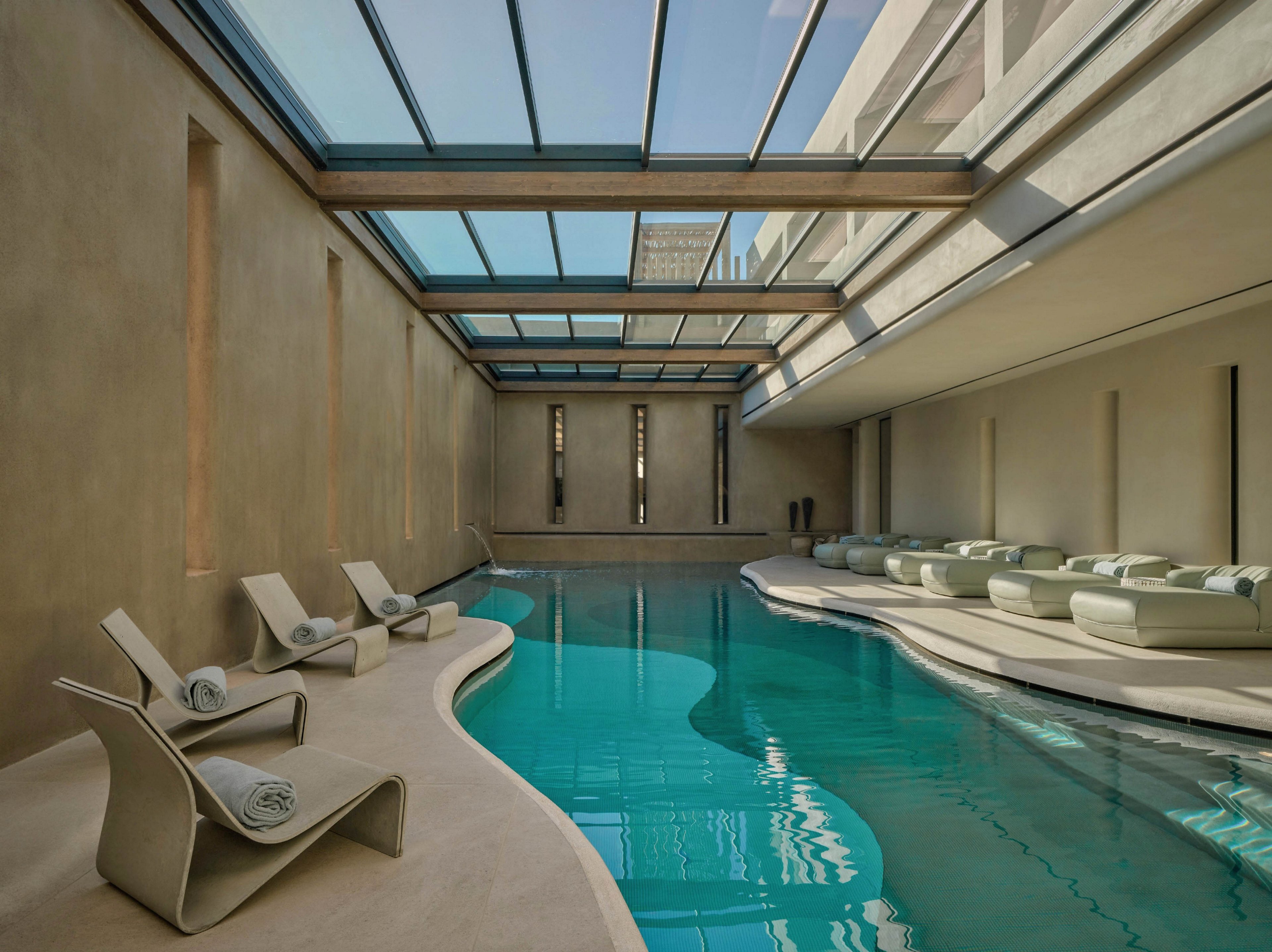 A contemporary indoor swimming pool is surrounded by sculptural lounge chairs and cushioned daybeds. The space features a long, narrow pool with curved edges and calm turquoise water. Large skylights above allow natural light to flood the minimalist, beige-toned interior. The overall atmosphere is serene and luxurious, ideal for relaxation and wellness.