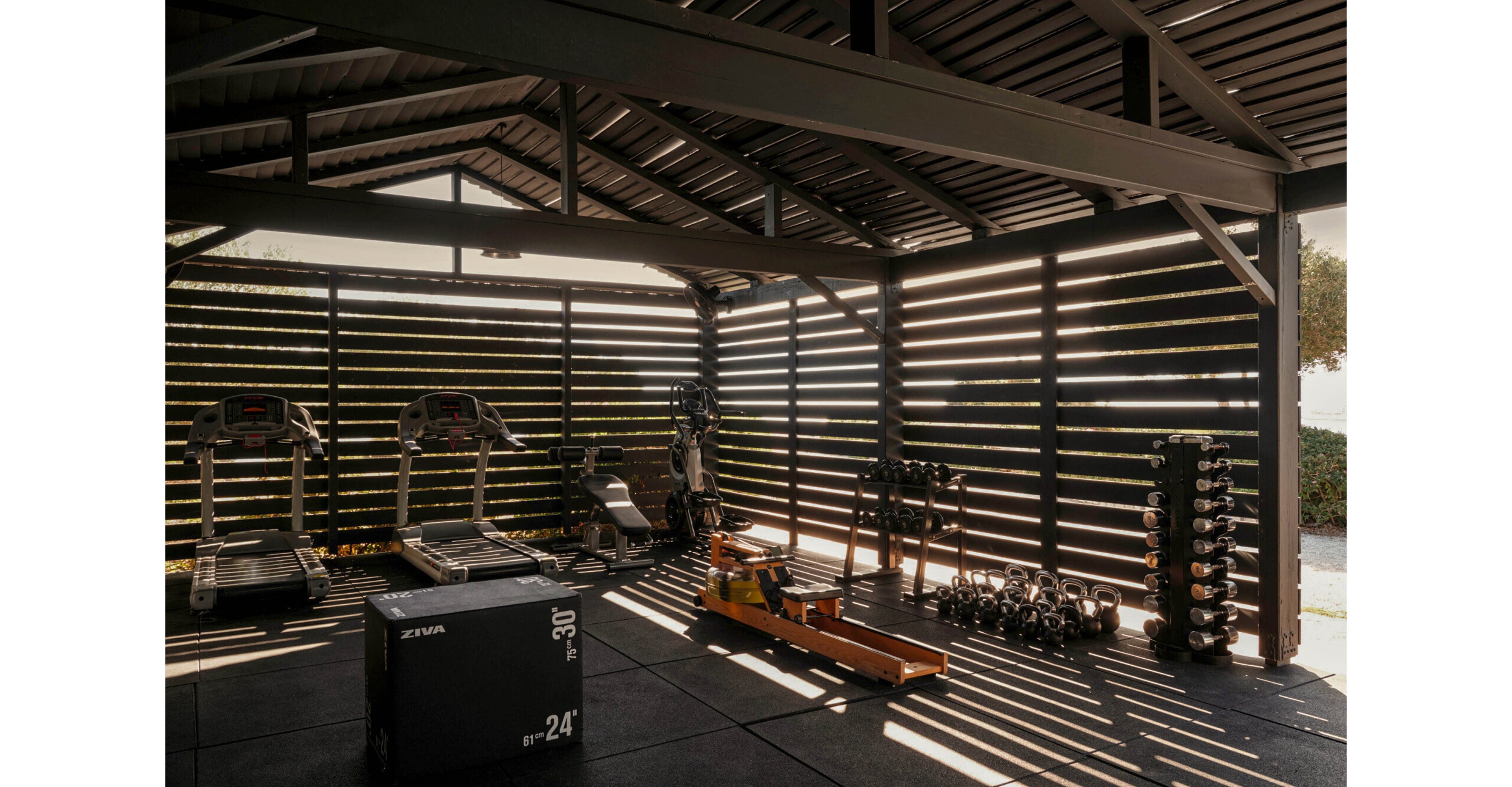 A covered outdoor gym features treadmills, weight benches, and free weights arranged neatly under a slatted roof. Sunlight streams through the horizontal panels, casting striped shadows across the floor and equipment. A plyometric box marked with visible text sits in the foreground, while dumbbells and kettlebells are organized on racks to the side. The space feels modern, functional, and designed for serious home workouts.