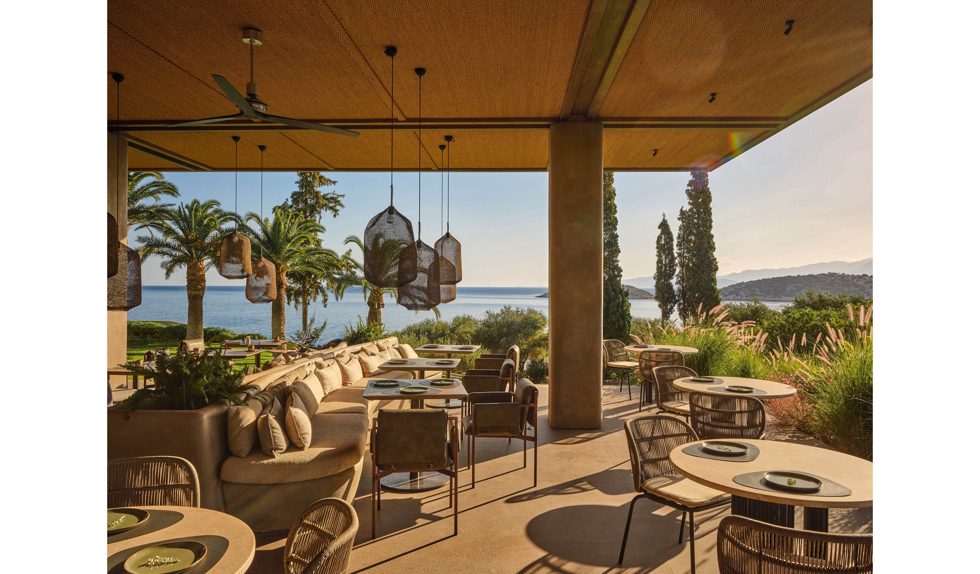 An elegant open-air restaurant terrace overlooks a calm sea and distant hills under warm evening light. Round tables with woven chairs and cushioned lounge seating create a relaxed, upscale dining atmosphere. Decorative hanging lanterns and ceiling fans add visual interest beneath the covered patio. Lush greenery, palm trees, and tall cypress trees frame the scenic waterfront view.