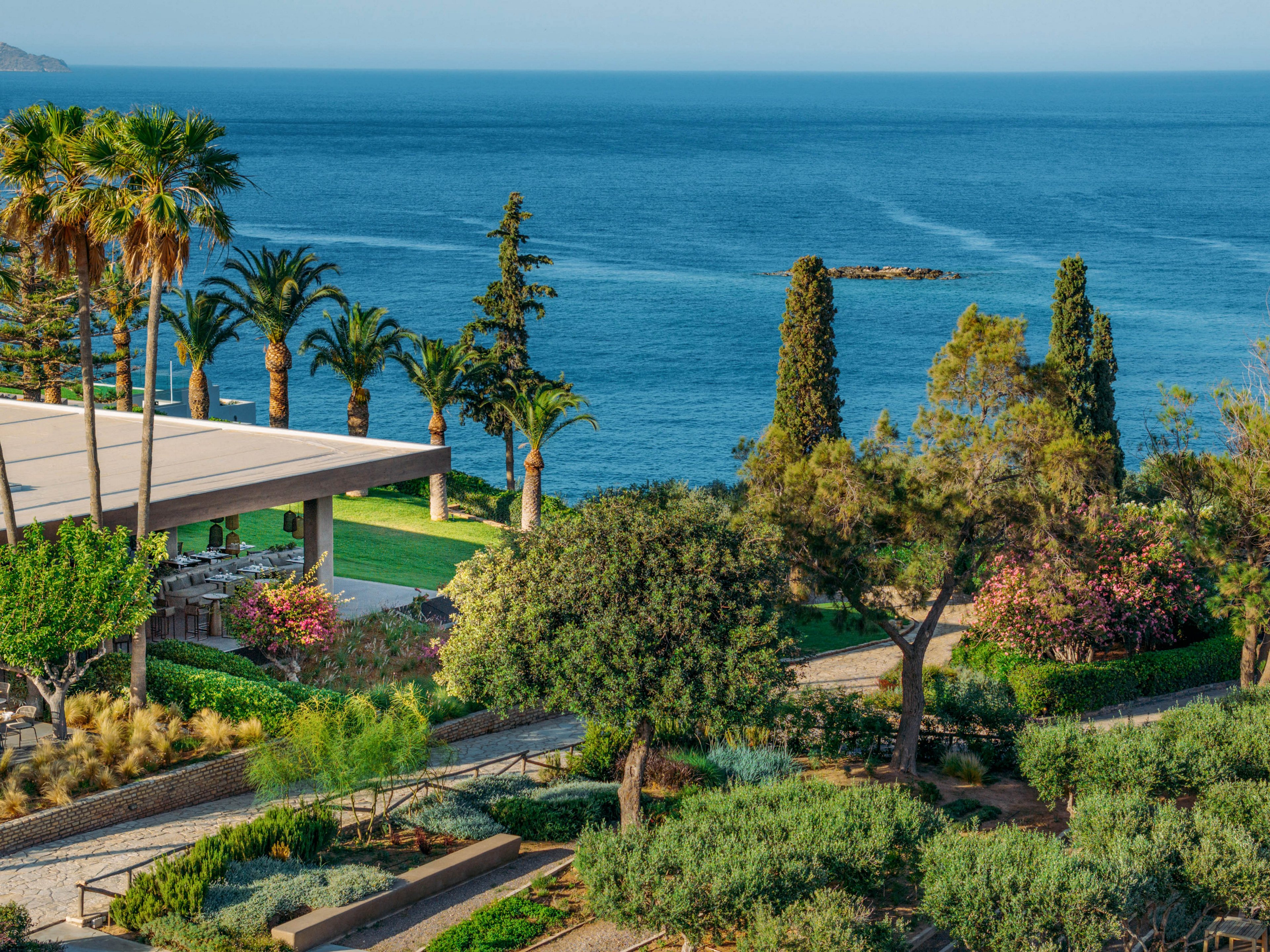 A landscaped coastal resort garden stretches toward a calm blue sea. Palm trees, shrubs, and flowering plants line stone paths and terraces around a modern open-air building. The scene is sunlit, with vibrant greenery contrasting against the deep blue water and clear sky. The overall atmosphere is tranquil and inviting, suggesting a relaxing vacation or retreat destination.