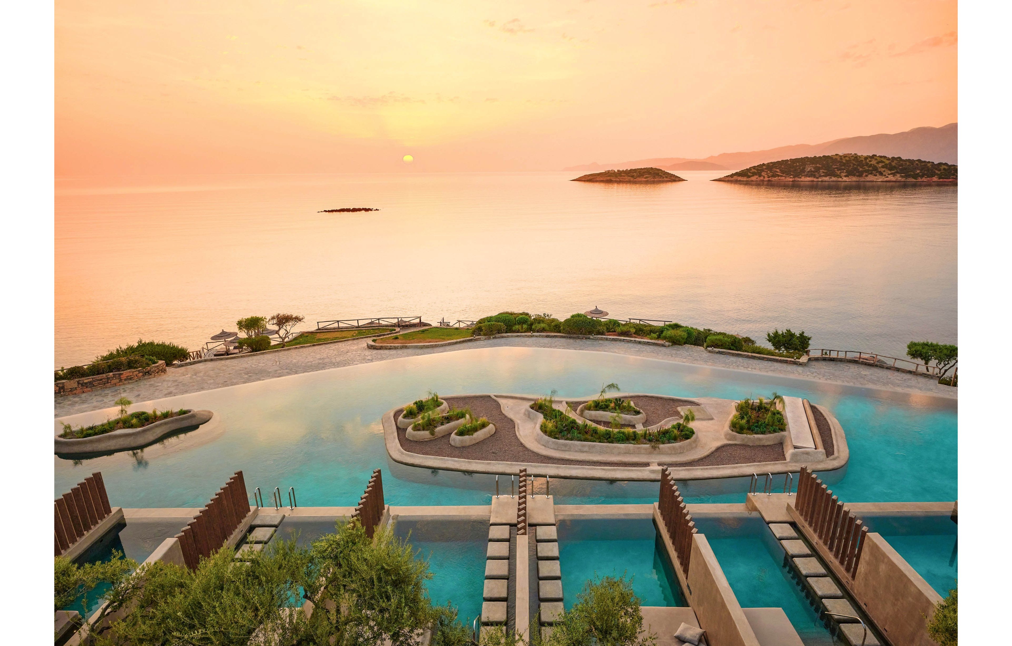 An expansive infinity-style pool overlooks a calm sea at sunset, with small islands visible on the horizon. Tiered swimming lanes and landscaped terraces create a geometric pattern leading toward the water. Soft orange and pink sky tones reflect on the surface, enhancing the tranquil atmosphere. Lush greenery and modern sunbathing areas suggest a high-end coastal resort setting.