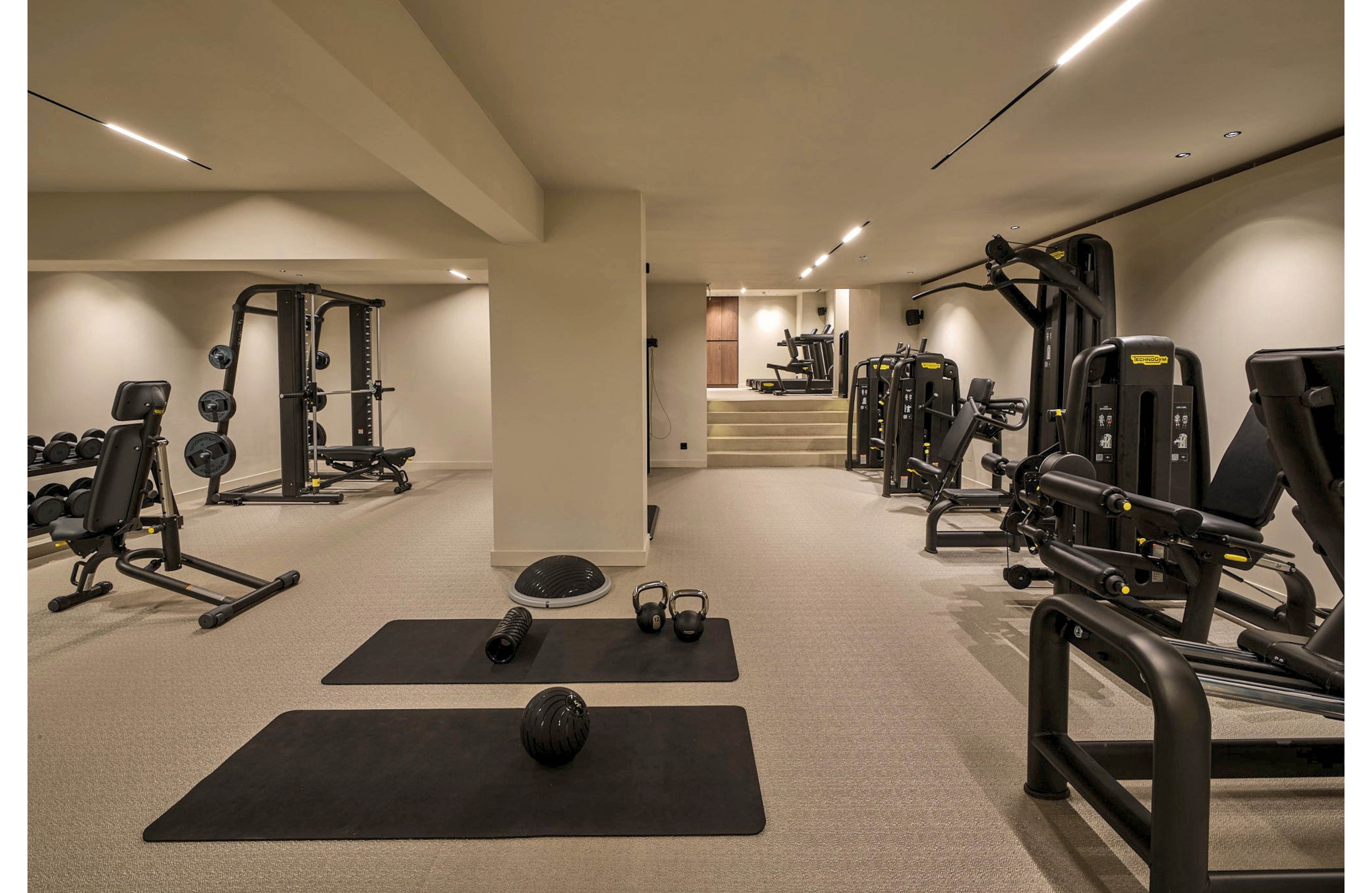 A spacious, modern indoor gym features multiple strength training machines and free weights arranged neatly across the room. Two black exercise mats lie in the foreground with kettlebells, a medicine ball, and a balance dome placed on the floor. The neutral color palette, recessed ceiling lights, and clean design create a calm, professional workout environment. In the background, additional fitness machines and a small raised platform are visible, emphasizing the variety of training options.