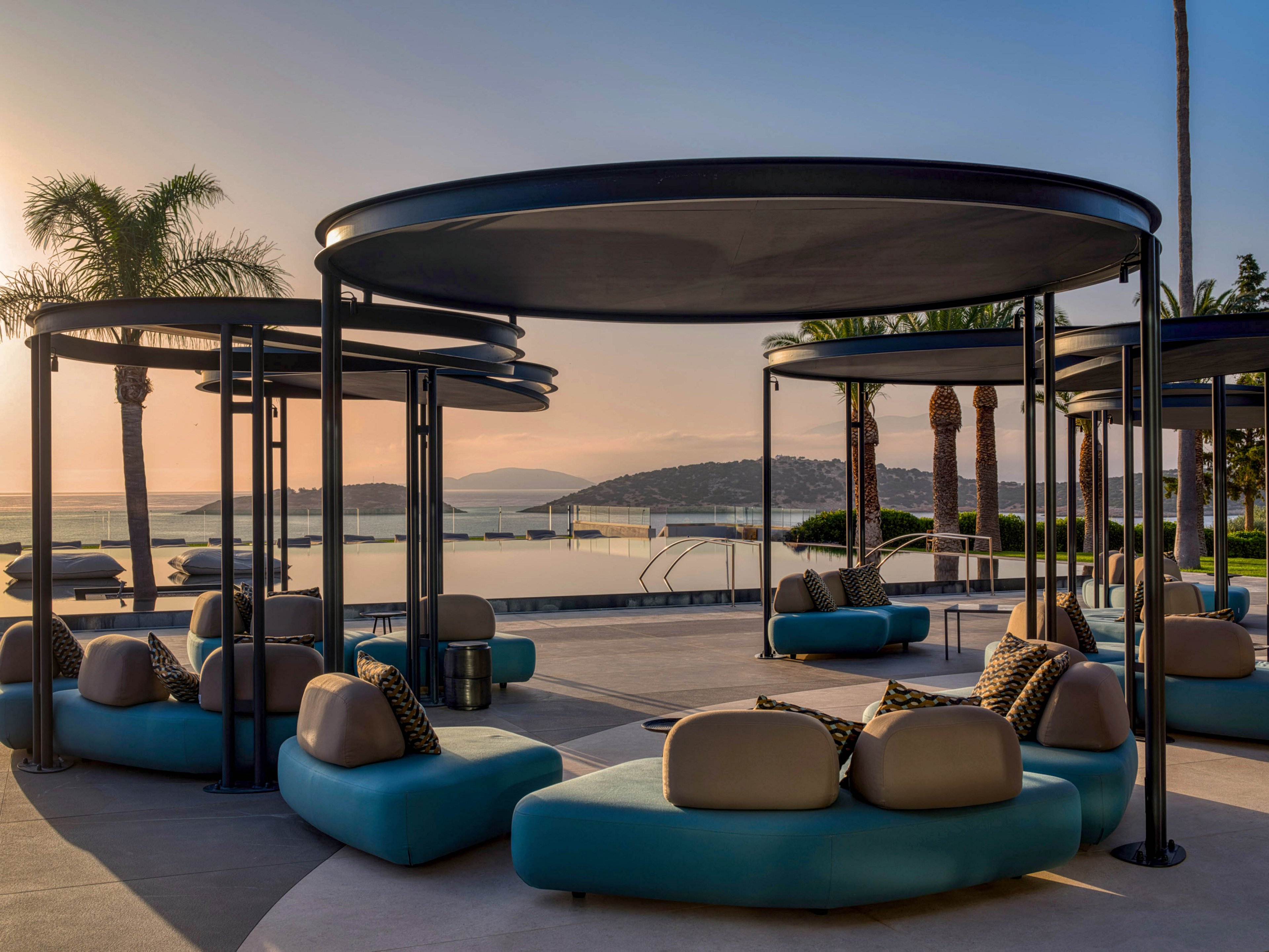 Contemporary outdoor lounge cabanas sit beside a calm waterfront at sunset. Curved turquoise seating with neutral cushions is arranged under circular metal canopies, creating a stylish relaxation area. Palm trees and an infinity-style pool frame the view toward distant hills and the sea. The warm evening light enhances the tranquil, luxurious resort atmosphere.