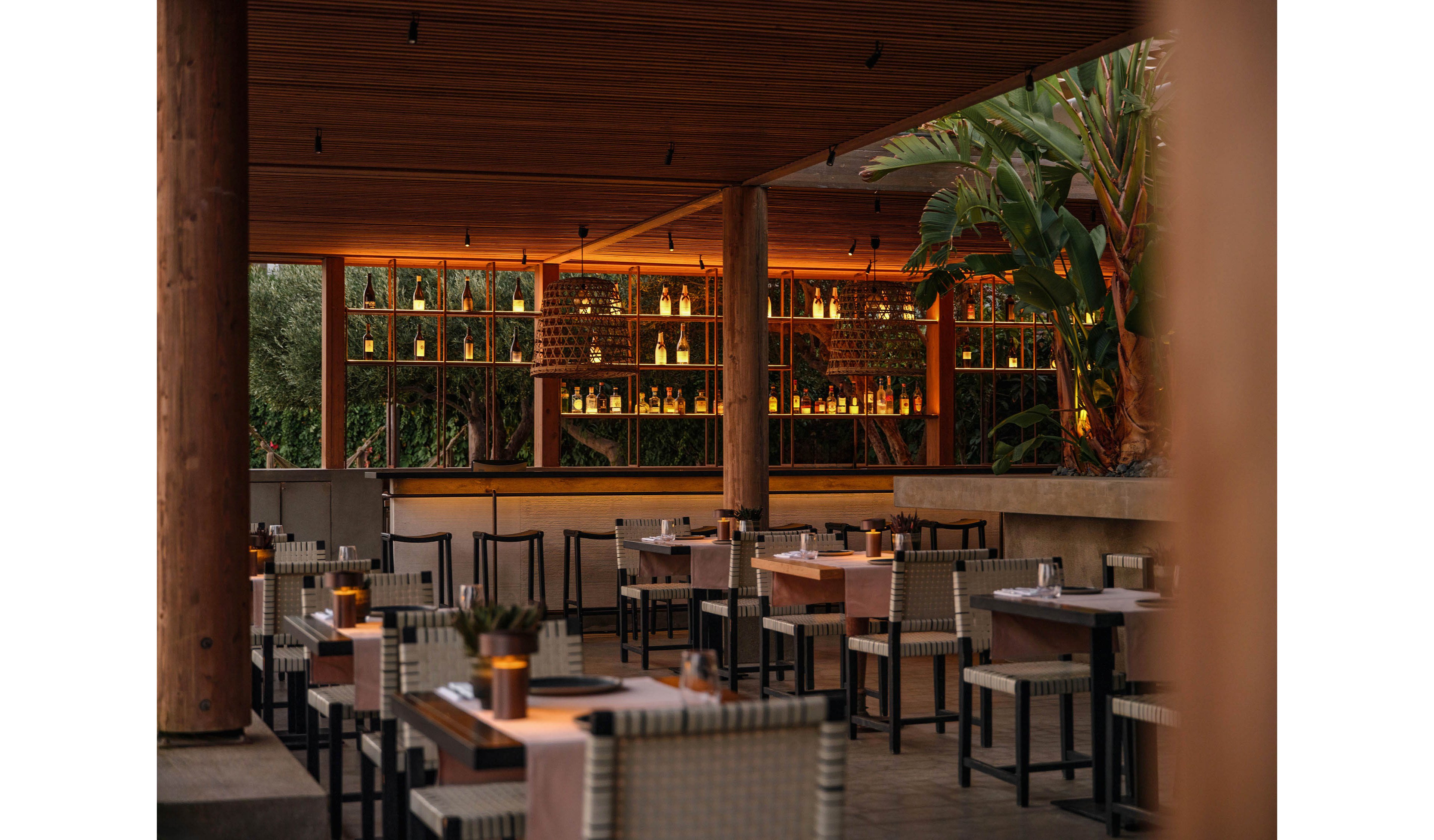 An open-air bar and dining area is shown at dusk, featuring neatly arranged tables and chairs. Warm ambient lighting illuminates the bar shelves filled with bottles, creating a cozy atmosphere. Wooden columns, a slatted ceiling, and lush tropical plants frame the space. The scene suggests a relaxed, upscale restaurant or lounge ready to welcome guests.