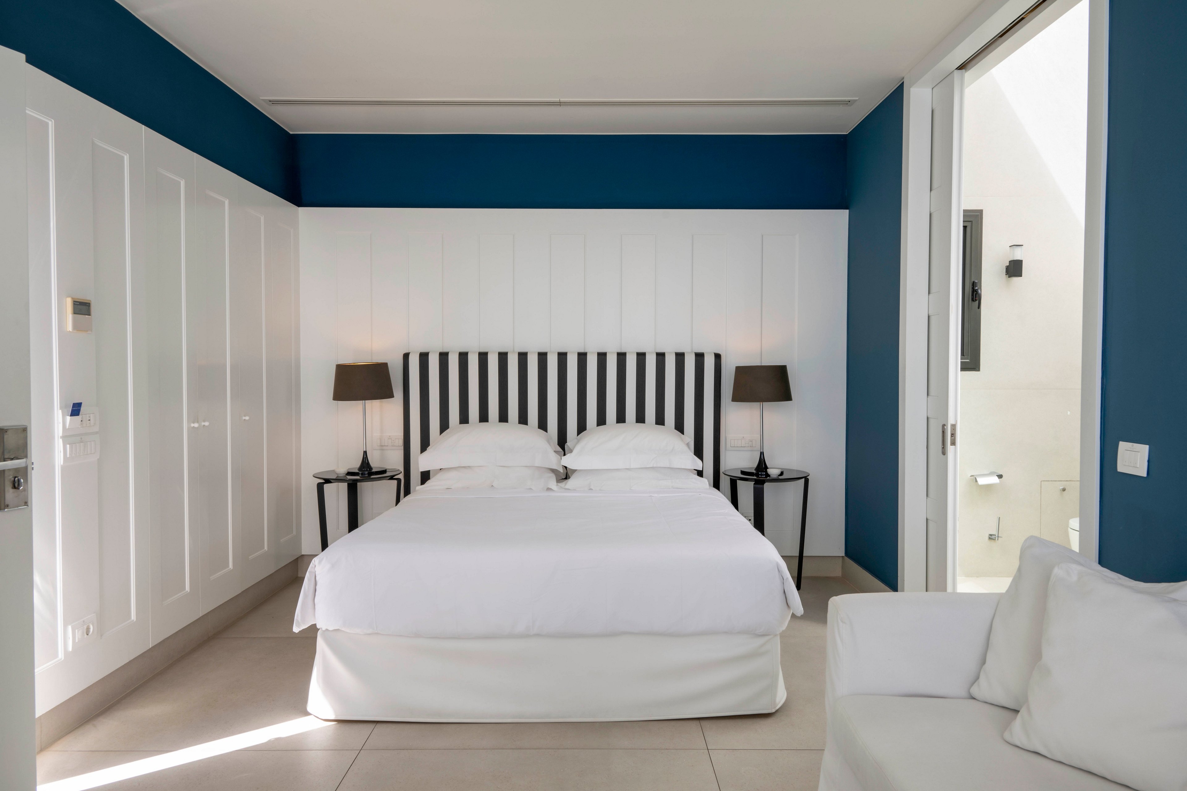 A neatly made double bed with a striped headboard stands centered in a modern hotel bedroom. The room features white paneled walls contrasted by a deep blue accent wall and light tiled flooring. Two bedside tables with matching dark lamps flank the bed, while a white sofa sits in the foreground. An open doorway on the right leads to a bright adjoining space, enhancing the airy, minimalist feel of the interior.