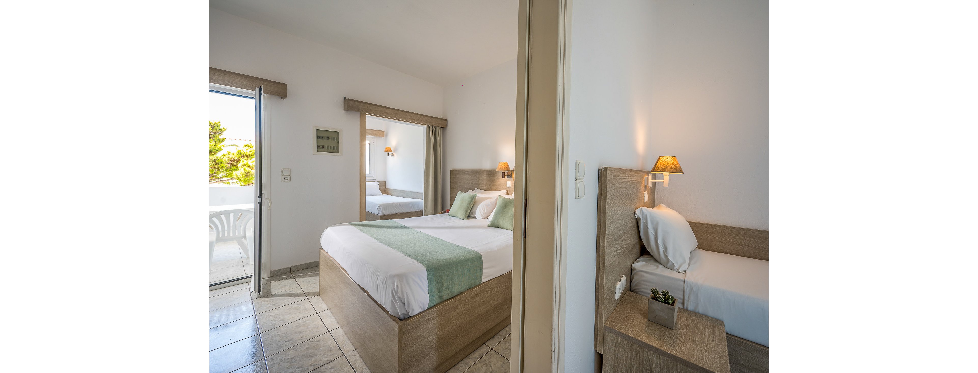 A modern hotel room featuring a double bed with light green accents and a single bed in an adjacent area. The setting includes a balcony with outdoor seating and natural light streaming in. Warm lighting and neutral tones create a relaxing atmosphere.