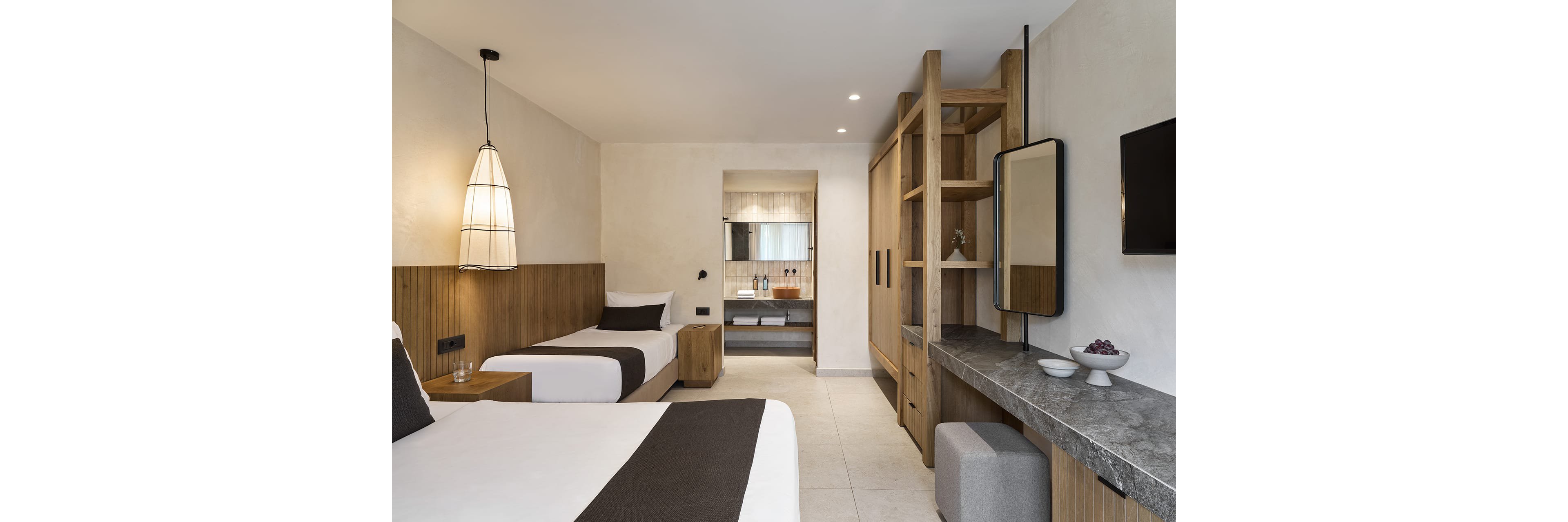 Contemporary hotel room featuring two single beds with white linens and dark throws. The space includes a built-in wooden headboard, open wardrobe, and a stone-topped desk with a wall mirror. Warm pendant lighting and neutral walls create a calm, minimalist atmosphere. In the background, an open doorway reveals the bathroom area with a sink and toiletries.