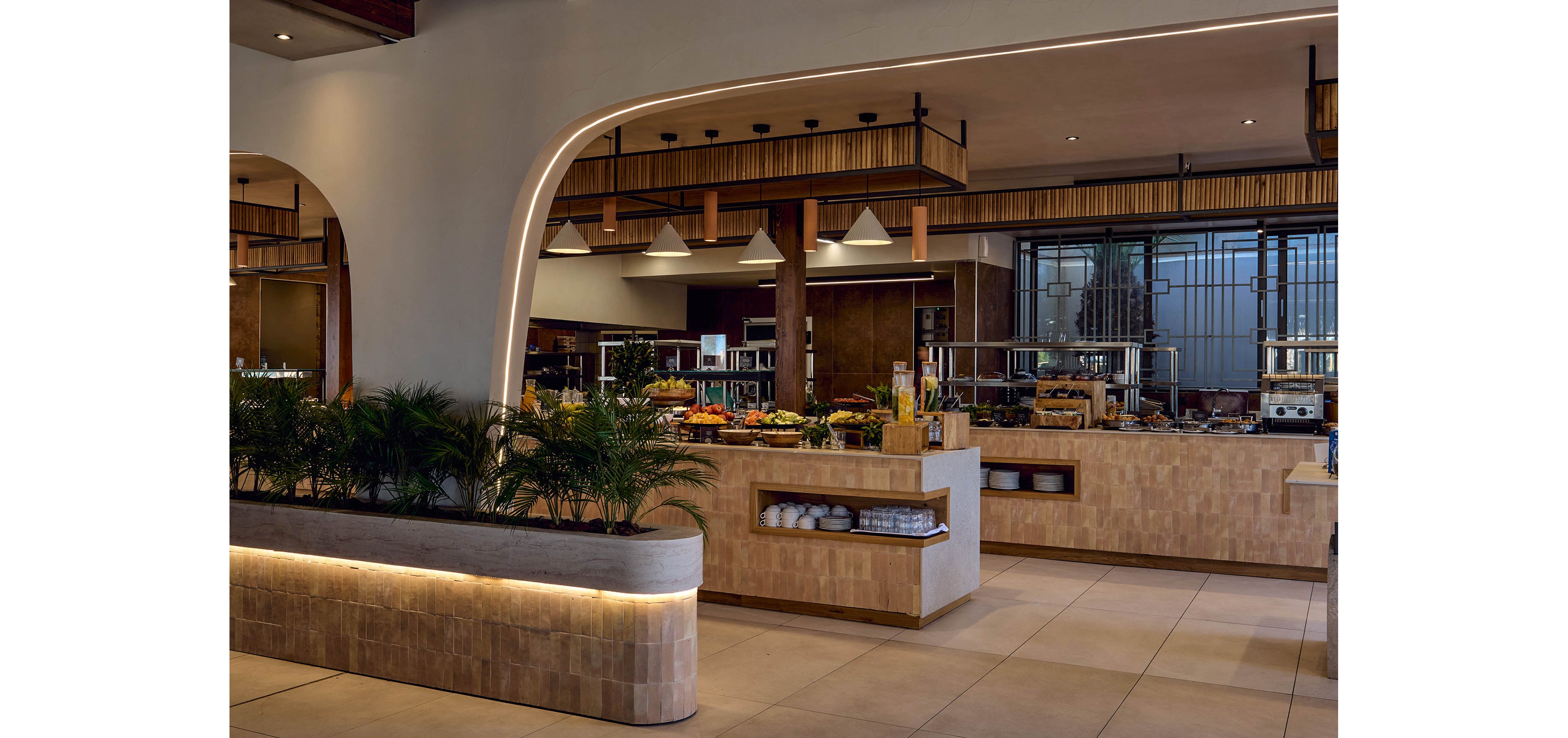 Contemporary buffet counters are arranged in a spacious restaurant interior with warm lighting. Various dishes and serving platters are displayed behind glass and on open surfaces, suggesting a self-service dining area. Architectural arches, wood accents, and tiled surfaces create a stylish, upscale atmosphere. Potted plants and clean, uncluttered floors contribute to a relaxed, inviting environment.