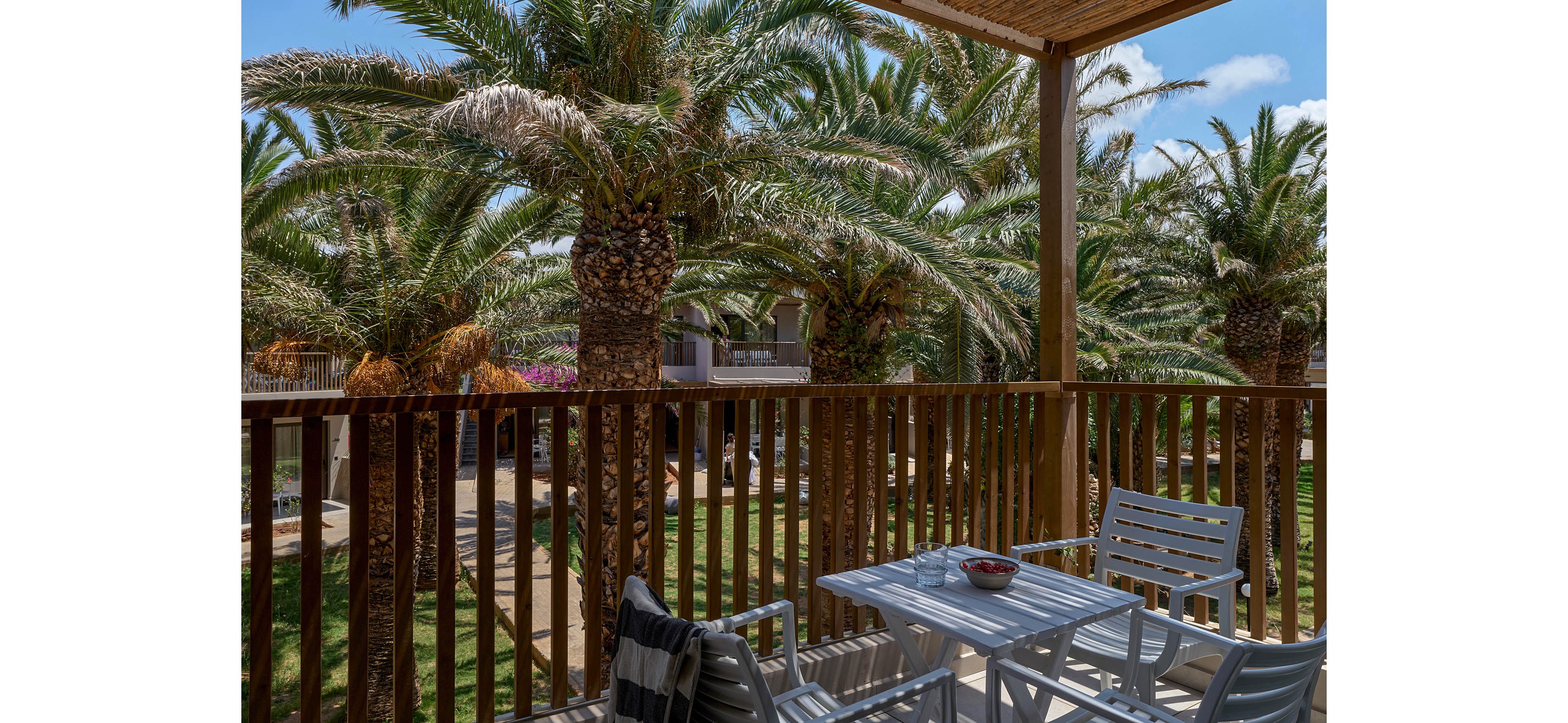 An outdoor balcony features a small white table and chairs overlooking a lush garden of tall palm trees. Wooden railings frame the view of the resort buildings and landscaped pathways below. A striped towel or blanket rests on one chair, and a small bowl sits on the table, suggesting a relaxed vacation setting. Bright daylight and blue sky create a warm, inviting atmosphere.