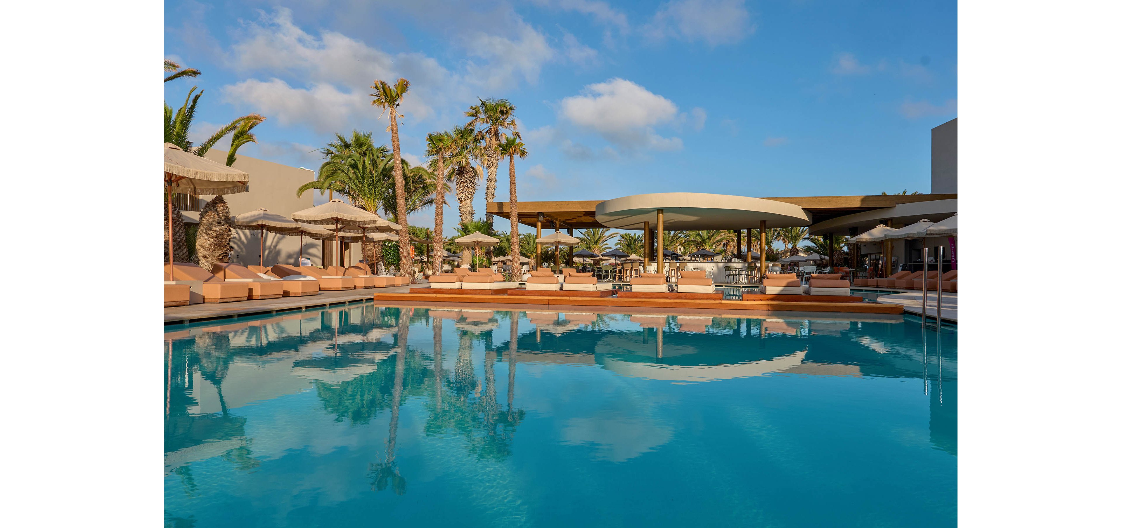 A large turquoise swimming pool reflects palm trees and a clear blue sky at a modern resort. Beige cushioned sun loungers with umbrellas line the poolside, creating a relaxed vacation atmosphere. Contemporary open-air structures and seating areas are visible in the background, suggesting a stylish outdoor bar or lounge. The scene is calm and inviting, ideal for leisure, sunbathing, and warm-weather travel.
