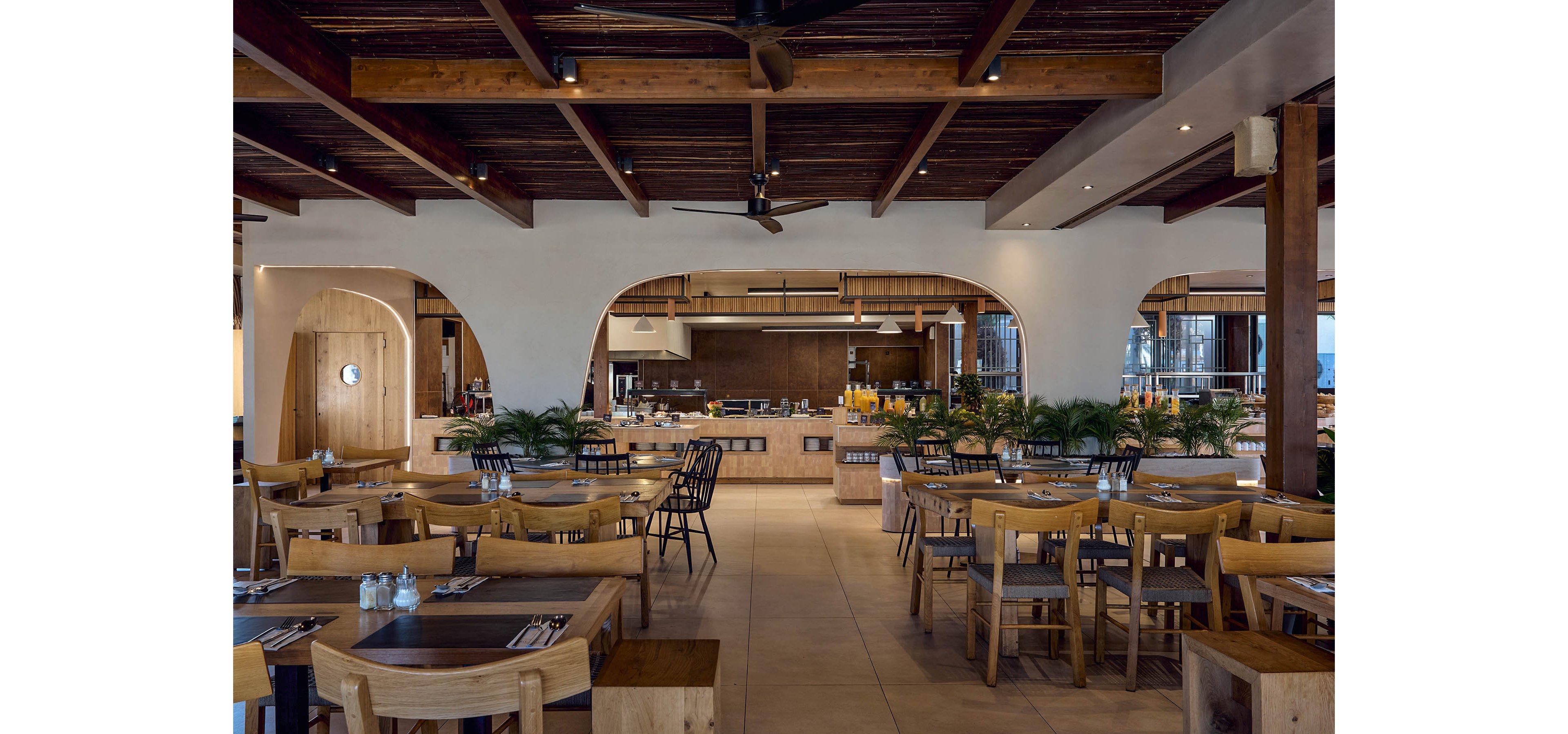 Spacious restaurant interior features wooden tables and chairs arranged neatly in an open-concept dining area. The setting includes a central buffet or serving counter with drinks and food displays in the background. Warm wood tones, white walls, and arched openings create a contemporary yet cozy atmosphere. Potted green plants add freshness and contrast to the neutral color palette, while ceiling lights provide soft, even illumination.