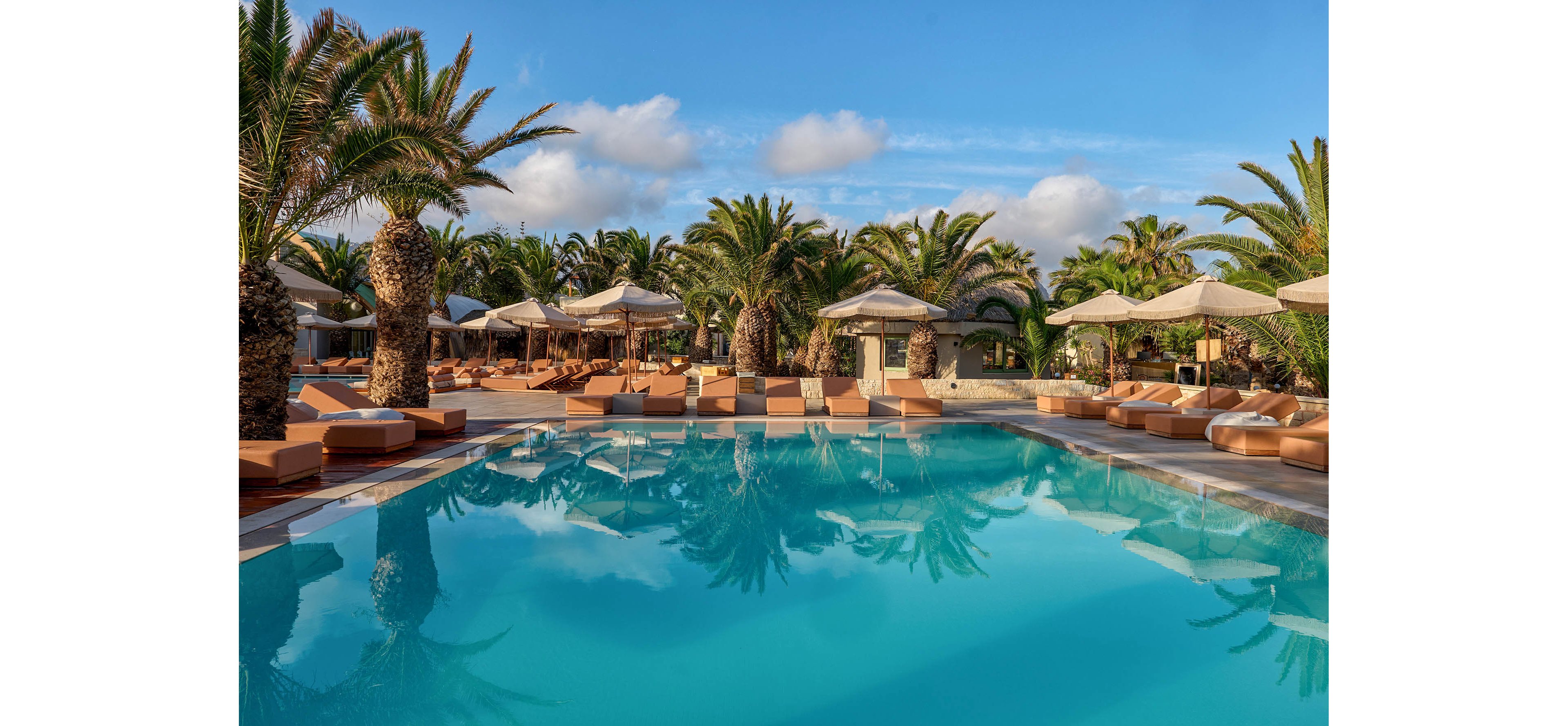 A tranquil resort swimming pool is surrounded by tall palm trees and cushioned lounge chairs. Beige umbrellas line the deck, creating shaded relaxation areas around the water. The clear blue pool reflects the sky and scattered clouds, enhancing the serene atmosphere. Warm tones of the seating contrast with the cool blue water, suggesting a peaceful vacation setting.