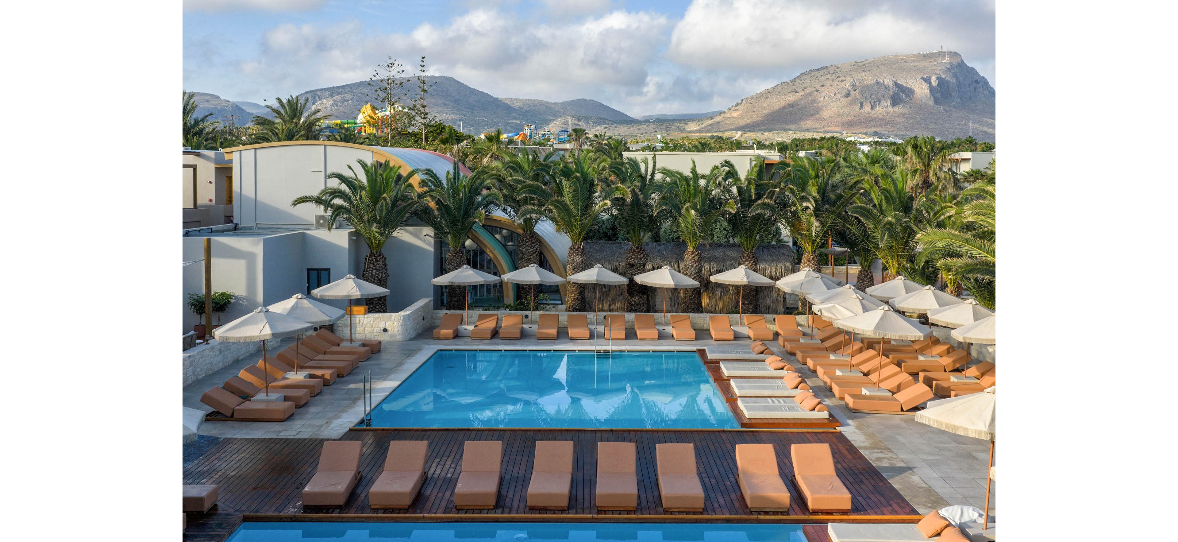 A large resort swimming pool is surrounded by neatly arranged sun loungers and umbrellas. The scene is framed by tall palm trees and low modern buildings in the background. Distant mountains rise under a partly cloudy sky, adding a scenic backdrop. The overall atmosphere suggests a calm, sunny vacation setting at a tropical or Mediterranean hotel.