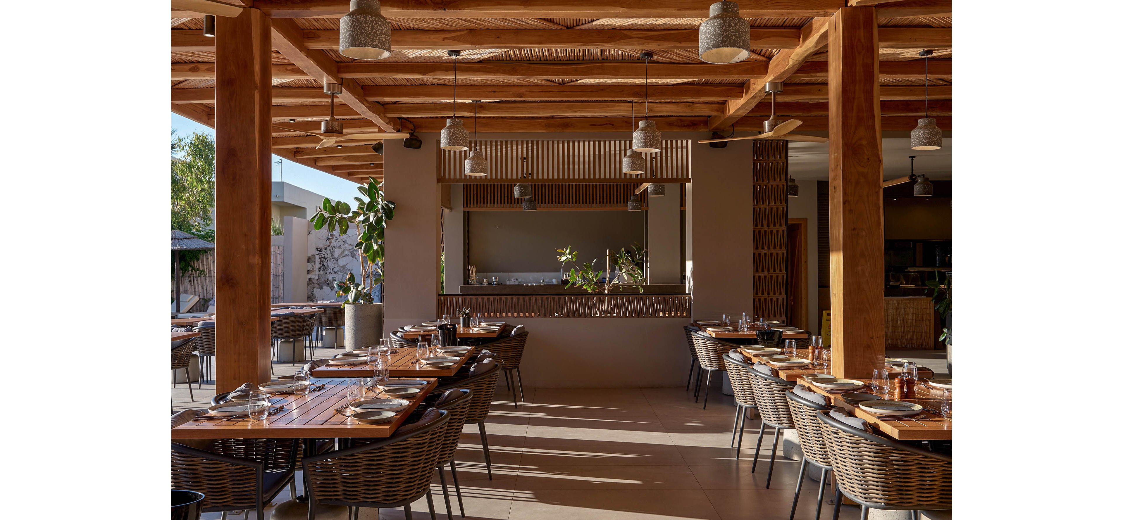 An inviting open-air restaurant features neatly arranged wooden tables and woven chairs under a warm timber ceiling. Sunlight streams through the space, highlighting table settings with plates, glasses, and cutlery ready for guests. Potted plants and natural materials create a relaxed, contemporary atmosphere. The overall scene feels calm and welcoming, ideal for casual dining or social gatherings.