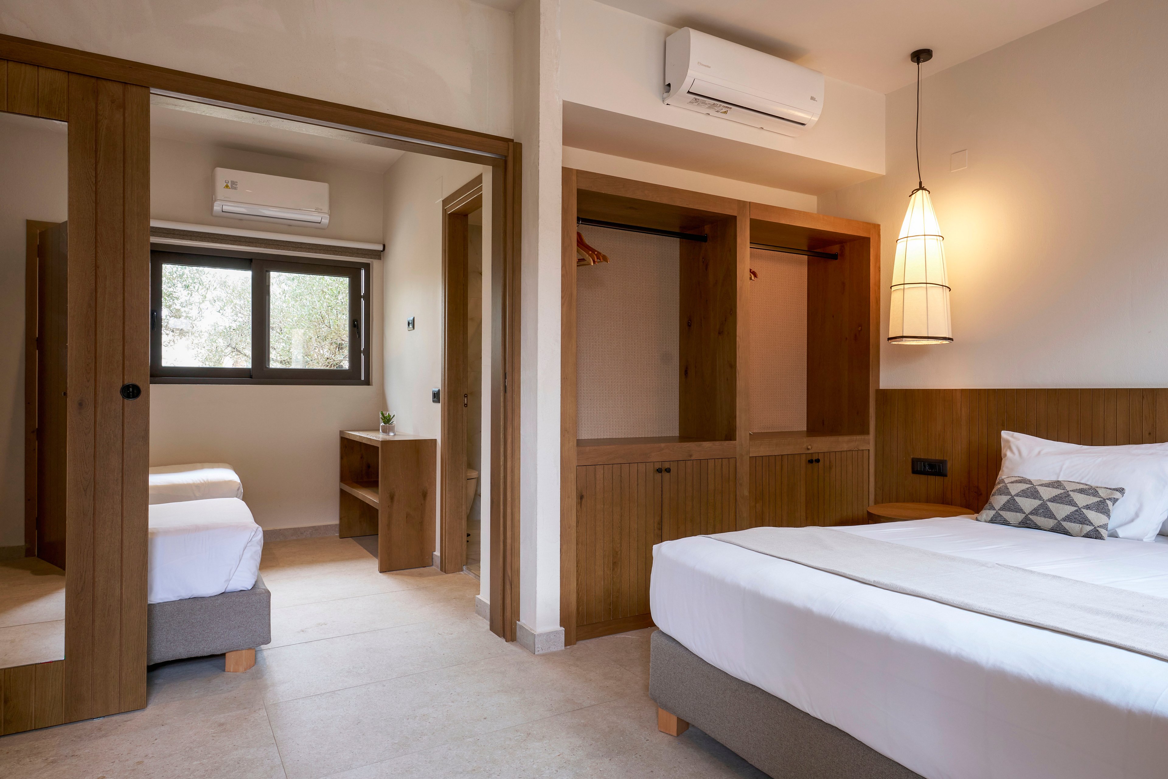 A contemporary hotel room features a neatly made double bed with a patterned cushion and wooden headboard. Built-in wooden wardrobes and an open closet provide storage along the wall. An adjoining room with twin beds is visible through a doorway, along with a wooden desk beneath a window. Neutral tones, soft lighting, and a hanging pendant lamp create a calm, minimalist atmosphere. A wall-mounted air conditioning unit is installed above the doorway for climate control.