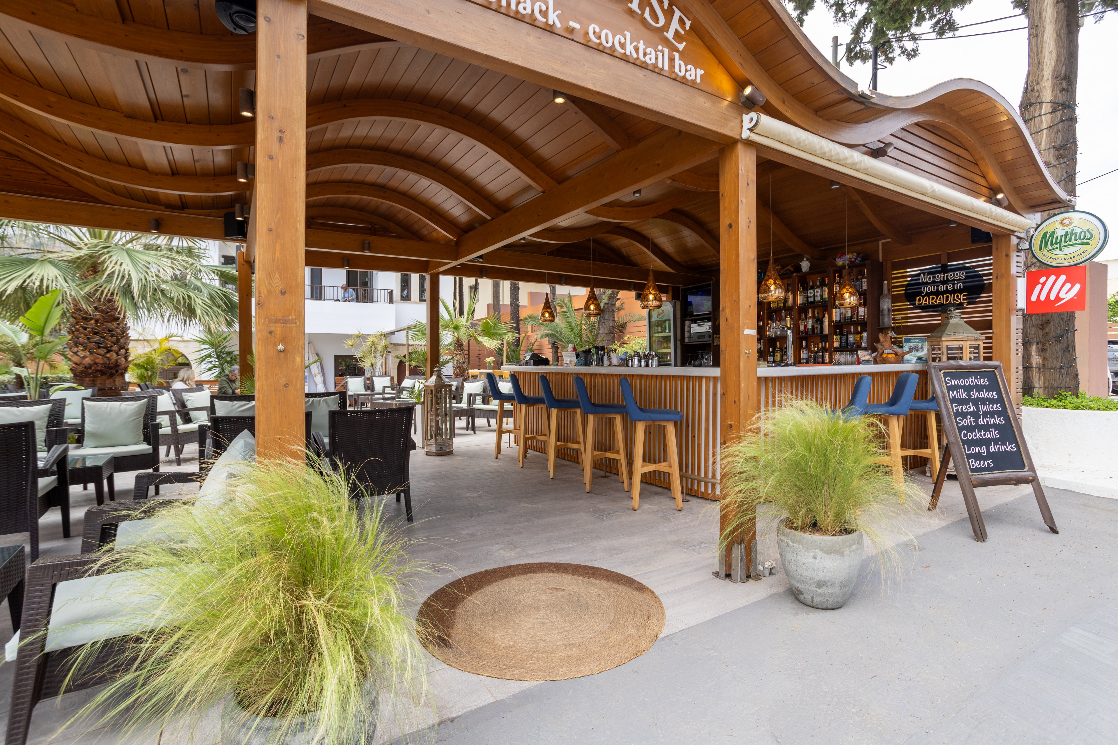 An open air wooden bar serves as a beach snack and cocktail bar with high stools lined along the counter. The setting features a curved timber roof, tropical plants, and outdoor seating with tables and chairs. Bottles and glasses hang behind the bar, while signs and menus are visible near the entrance. The relaxed atmosphere suggests a casual vacation or resort environment.